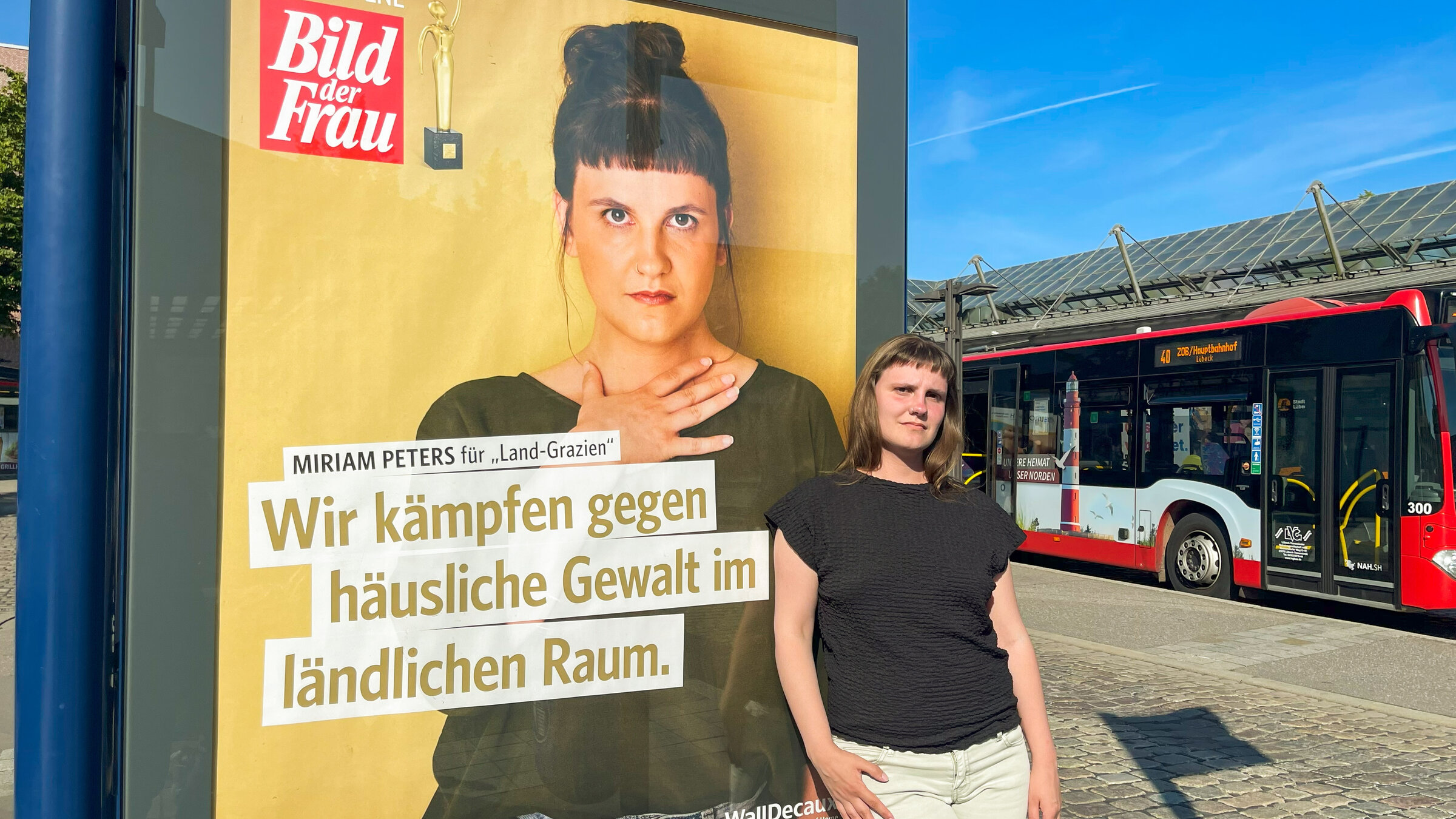 "Tatort Land: Rettung aus häuslicher Gewalt": Einem Werbeplakat hängt neben einer Bushaltestelle. Auf dem Plakat ist Miriam Peters als Preisträgerin von "Bild der Frau" zu sehen, darunter ist der Schriftzug "Wir kämpfen gegen häusliche Gewalt im ländlichen Raum". An der Plakatwand lehnt Miriam Peters und schaut in die Kamera. Im Hintergrund fährt gerade ein Bus vorbei.