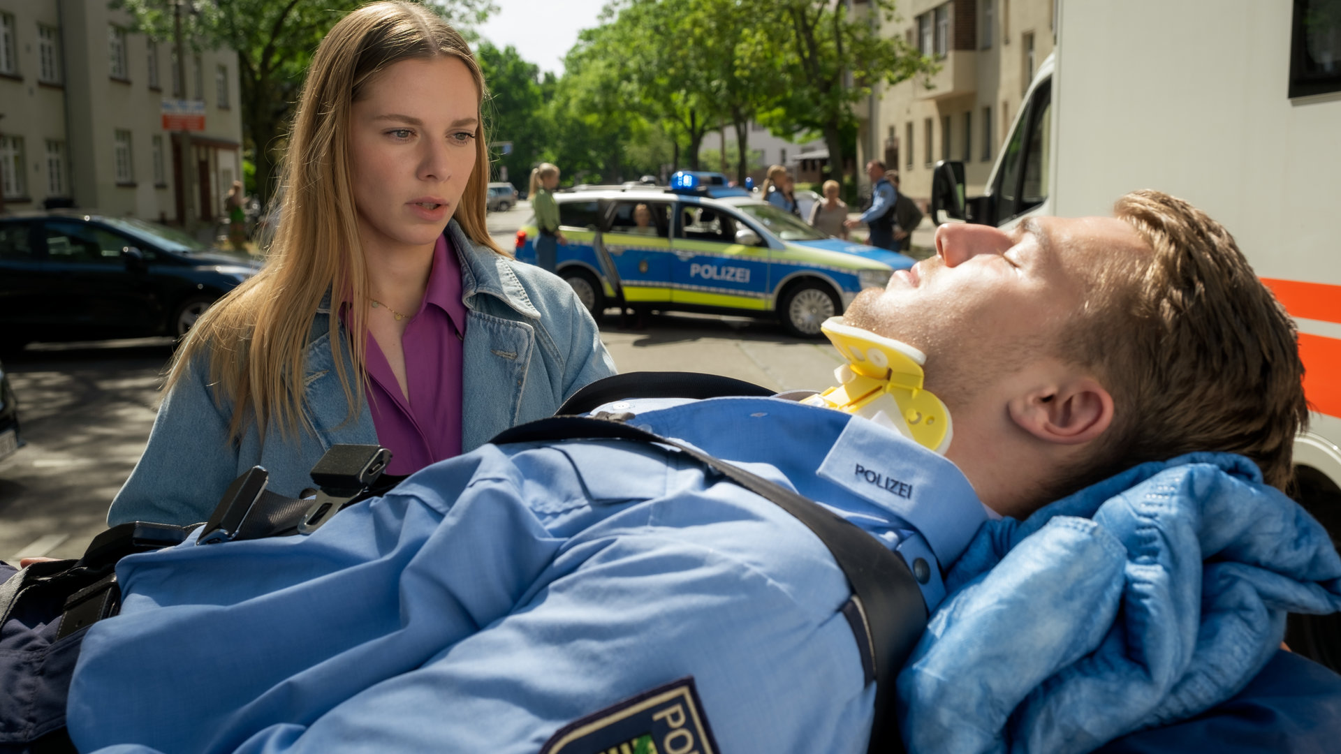 "Blutige Anfänger - Stiller Alarm": Phillip (Eric Cordes) liegt auf einer Patiententrage. Er hat die Augen geschlossen. Charlotte (Cheyenne Pahde) steht neben der Trage. Sie schaut ihn besorgt an. Im Hintergrund steht ein Notarztwagen und ein Streifenwagen.