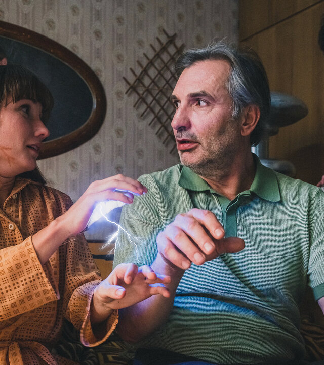 "We're on it, Comrades! - Liebling, du bist jetzt Witwe": Während Vojta (Jan Cina) im Hintergrund an einer Bedieneinheit seiner Geisterfalle hantiert, bekommt der auf dem Bett sitzende David (Jiří Macháček) von der neben ihm sitzenden Frau Kapounová (Eliška Křenková) einen elektrischen Schlag, der durch einen blauen Lichtbogen sichtbar wird.