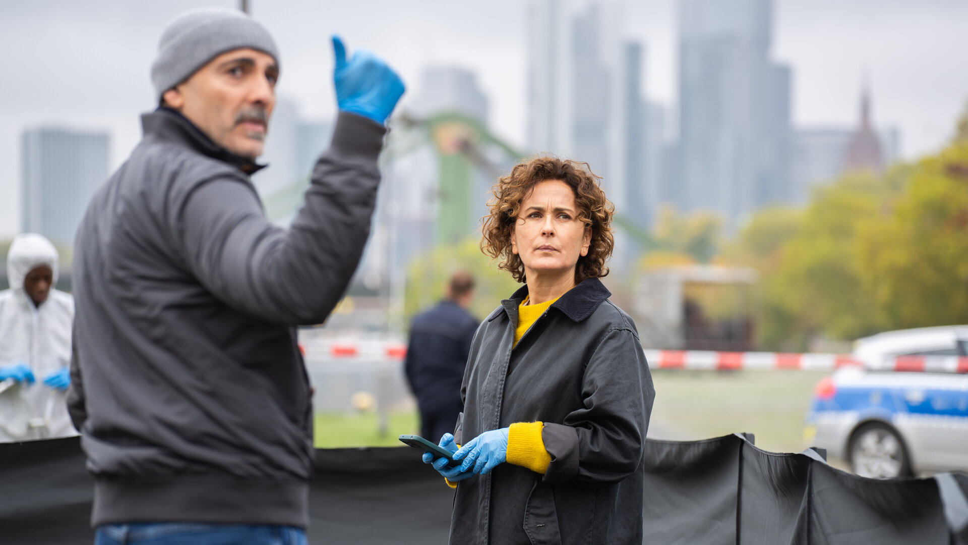 "Frankfurt Mord – Ein neues Leben": Die Ermittler Sarah Frankfurter (Marie-Lou Sellem) und Fêris Hahn (Billey Demirtaş) stehen im Vordergrund eines abgesperrten Tatorts am Mainufer. Beide tragen blaue Einweghandschuhe und blicken konzentriert in dieselbe Richtung. Hinter ihnen ist ein rot-weißes Absperrband gespannt. Dahinter parkt ein Polizeifahrzeug neben dem Einsatzkräfte (Komparsen), darunter eine Person in einem weißen Schutzanzug, vorbeigehen. Im Hintergrund liegt die Frankfurter Skyline vor grauem Himmel.