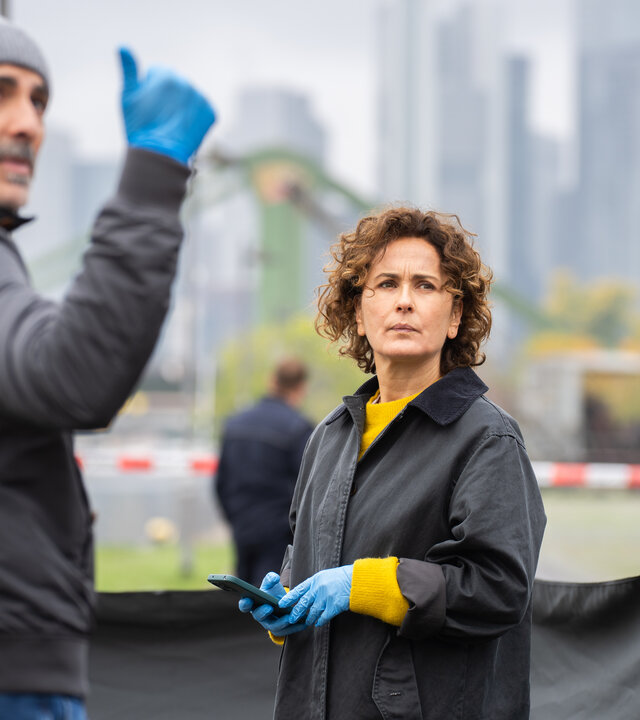 "Frankfurt Mord – Ein neues Leben": Die Ermittler Sarah Frankfurter (Marie-Lou Sellem) und Fêris Hahn (Billey Demirtaş) stehen im Vordergrund eines abgesperrten Tatorts am Mainufer. Beide tragen blaue Einweghandschuhe und blicken konzentriert in dieselbe Richtung. Hinter ihnen ist ein rot-weißes Absperrband gespannt. Dahinter parkt ein Polizeifahrzeug neben dem Einsatzkräfte (Komparsen), darunter eine Person in einem weißen Schutzanzug, vorbeigehen. Im Hintergrund liegt die Frankfurter Skyline vor grauem Himmel.