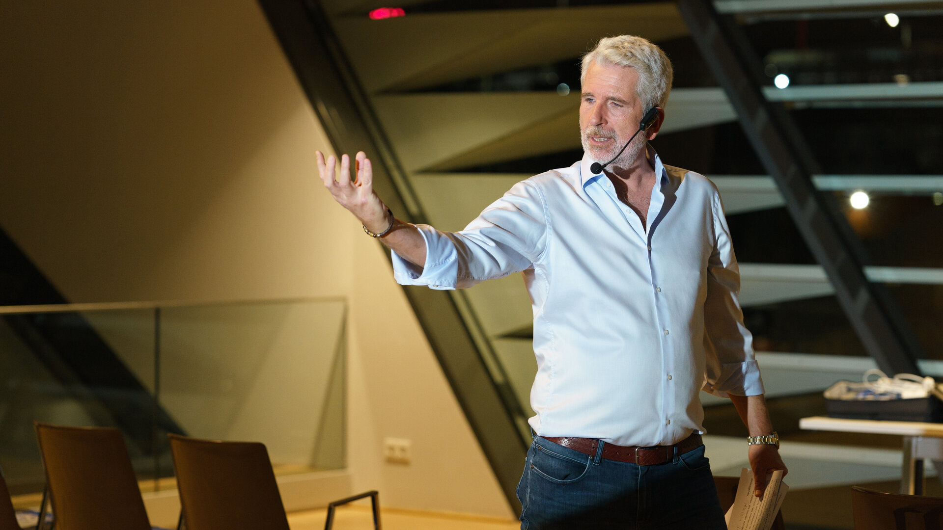 "SOKO Wien - Die 3 Säulen des Erfolgs": Mike Strasser (Dirk Stermann) steht breitbeinig im Konferenzsaal vor leeren Stühlen und hat seinen rechten Arm theatralisch erhoben. In der linken Hand hält er mehrere Moderationskarten. Mike Strasser trägt ein schwarzen Headset-Mikrofon.