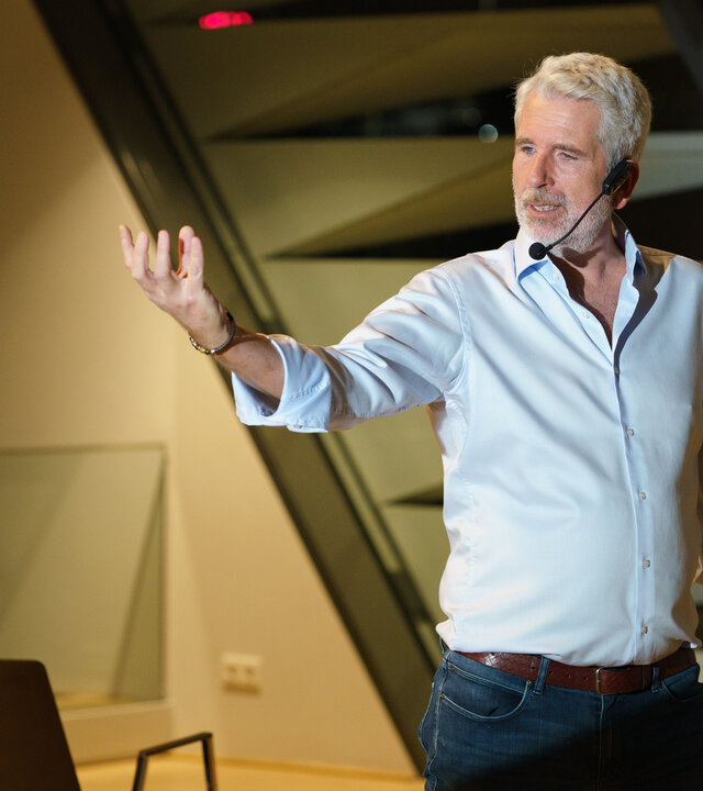 "SOKO Wien - Die 3 Säulen des Erfolgs": Mike Strasser (Dirk Stermann) steht breitbeinig im Konferenzsaal vor leeren Stühlen und hat seinen rechten Arm theatralisch erhoben. In der linken Hand hält er mehrere Moderationskarten. Mike Strasser trägt ein schwarzen Headset-Mikrofon.