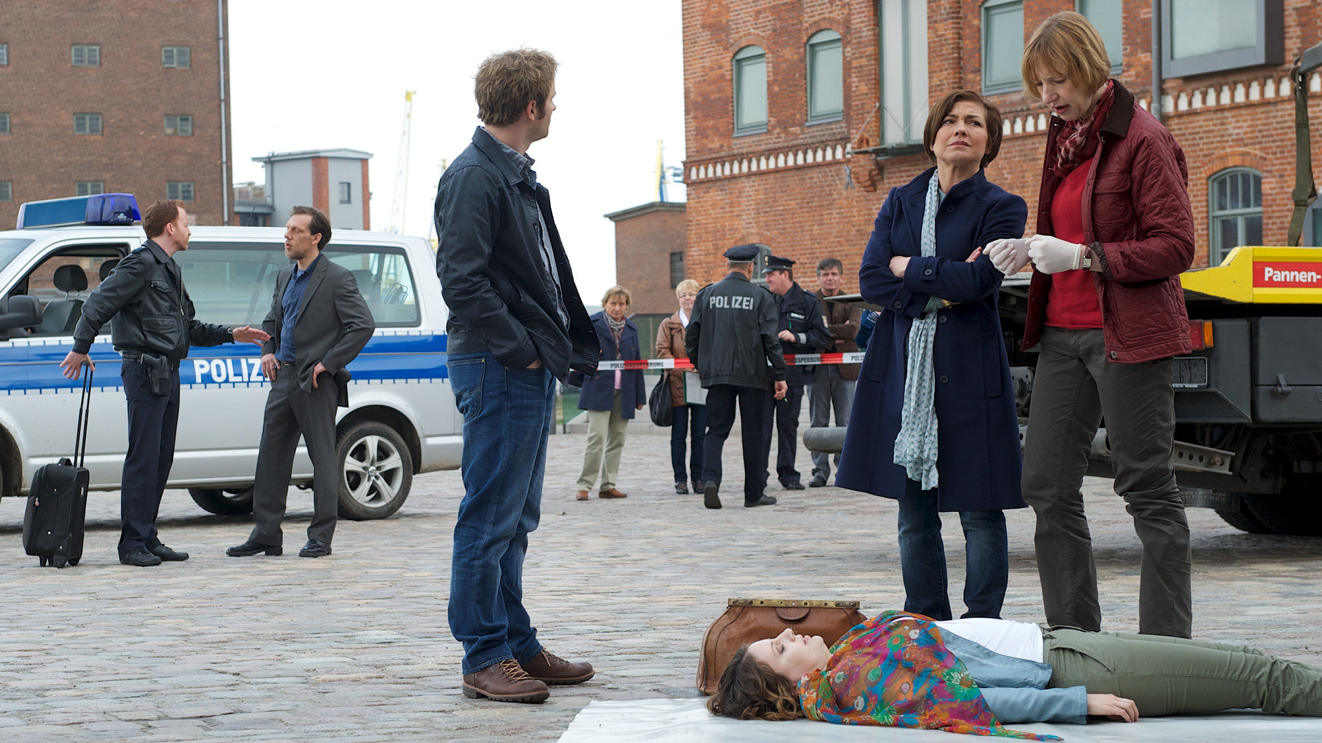 "SOKO Wismar - Sprachlos": Nils Theede (Jonas Laux), Katrin Börensen (Claudia Schmutzler) und Dr. Helene Sturbeck (Katharina Blaschke) besprechen am Tatort die ersten Erkenntnisse, während sie über der Leiche von Martina Opitz (Anja Margoni) stehen. Der Tatort selbst ist ein gepflasterter Innenhof. Im Hintergrund vom Tatort sind Leena Virtanen (Li Hagman) und Kai Timmermann (Mathias Junge).