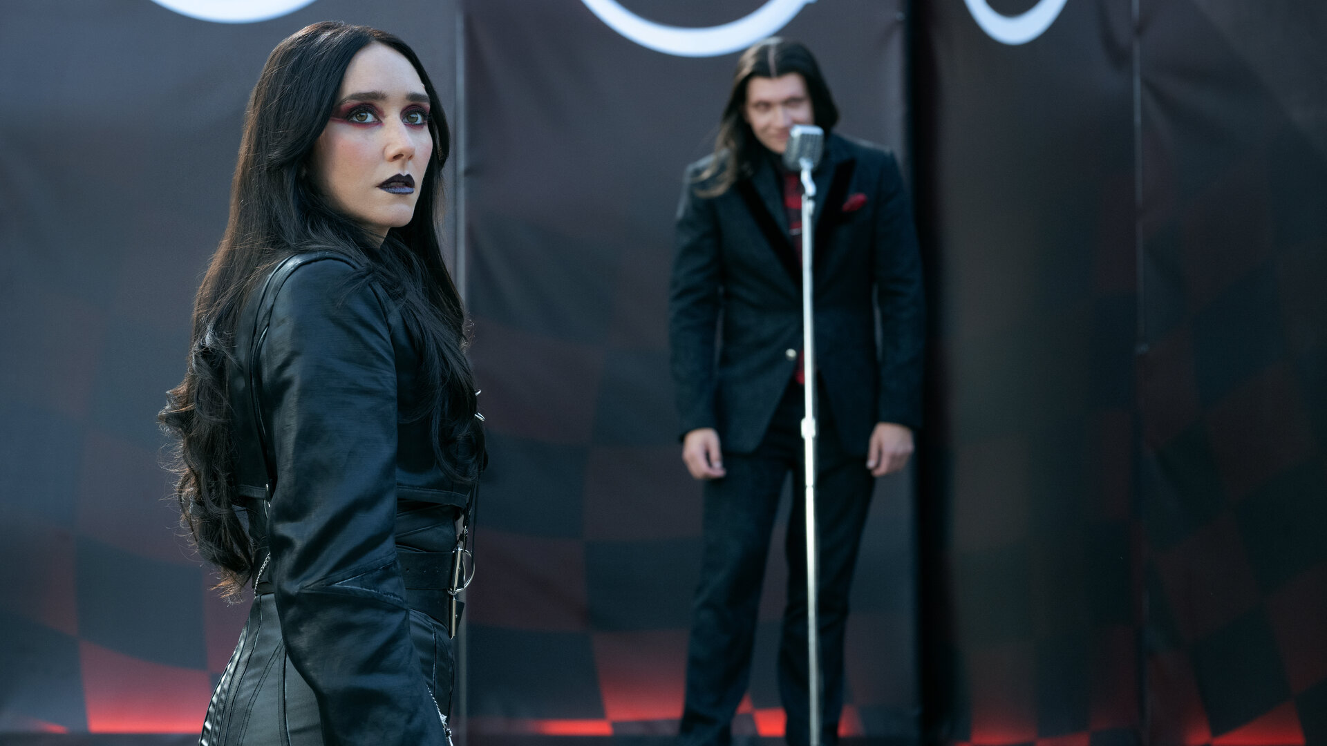 "Twisted Metal - MKAW1SH": Raven (Patty Guggenheim) steht im Publikumsbereich vor einer Bühne und blickt mit neutralem Ausdruck zur Seite. Hinter ihr steht Calypso (Anthony Carrigan) auf der Bühne an einem silbernen Mikrofon. Er nimmt eine leicht vorgebeugte Haltung ein und blickt erwartungsvoll zur Seite. Die Bühne wird von schwarzen Bannern mit roten Mustern und weißen Symbolen eingerahmt.
