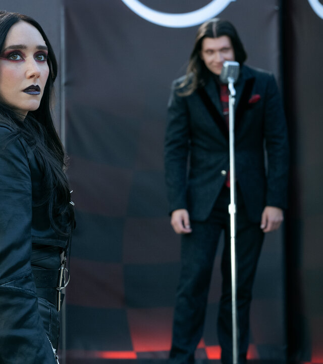 "Twisted Metal - MKAW1SH": Raven (Patty Guggenheim) steht im Publikumsbereich vor einer Bühne und blickt mit neutralem Ausdruck zur Seite. Hinter ihr steht Calypso (Anthony Carrigan) auf der Bühne an einem silbernen Mikrofon. Er nimmt eine leicht vorgebeugte Haltung ein und blickt erwartungsvoll zur Seite. Die Bühne wird von schwarzen Bannern mit roten Mustern und weißen Symbolen eingerahmt.