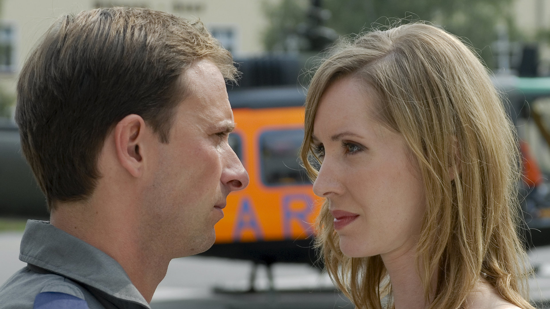 "Die Rettungsflieger - Entscheidungen": Außenaufnahme - Großaufnahme von Blank (Nicolas König) und seiner Freundin Juliane (Silvina Buchbauer), die beiden stehen dicht voreinander und sehen sich an. Im Hintergrund verschwommen ein Rettungshubschrauber.