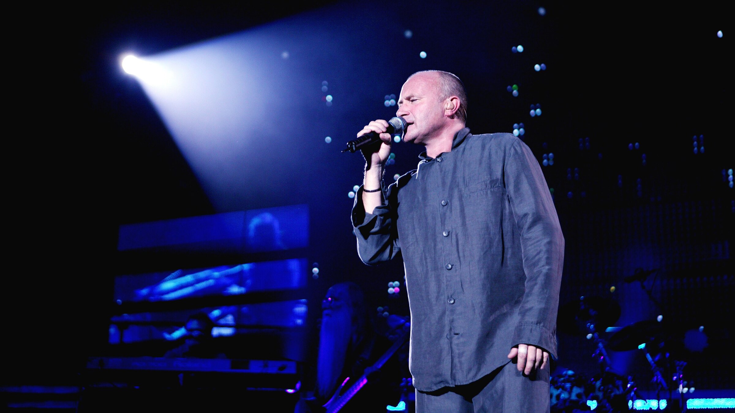 "Collins, Knopfler, Sting, Clapton, Elton John & Paul McCartney: Music for Montserrat": Der Musiker Phil Collins während eines Konzerts auf der Bühne. Er hält ein Mikrofon in der Hand und singt. Der Hintergrund ist in ein blau-schwarzes Licht getaucht.