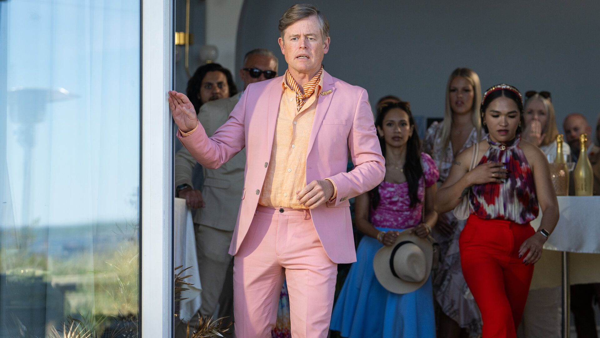 "Dan Sommerdahl - Tödliche Idylle: Miss Sunshine": Arne Lind (Caspar Phillipson) steht im Terrasseneingang eines Hotels und blickt entsetzt nach draußen. Seine rechte Hand greift zum offenen Schiebefenster. Er trägt einen Anzug in Rosa. Im Veranstaltungsraum hinter ihm blicken weitere festlich gekleidete Menschen (Komparsen) ebenso schockiert nach draußen.