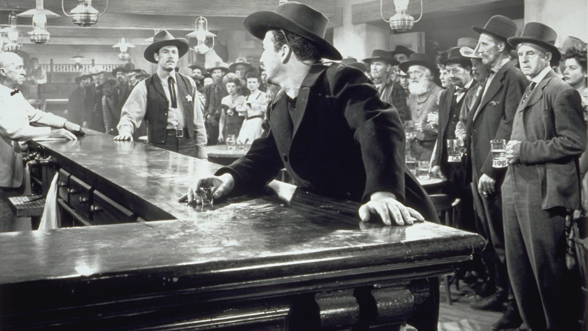 Bildbeschreibung „Faustrecht der Prärie“: In einem Saloon stehen sich zwei Männer am Tresen gegenüber, im Vordergrund stützt sich einer von beiden am Tresen ab. Im Hintergrund steht der andere, der einen Sheriffstern trägt. Die übrigen Gäste stehen rechts von ihnen  und verfolgen die Begegnung.