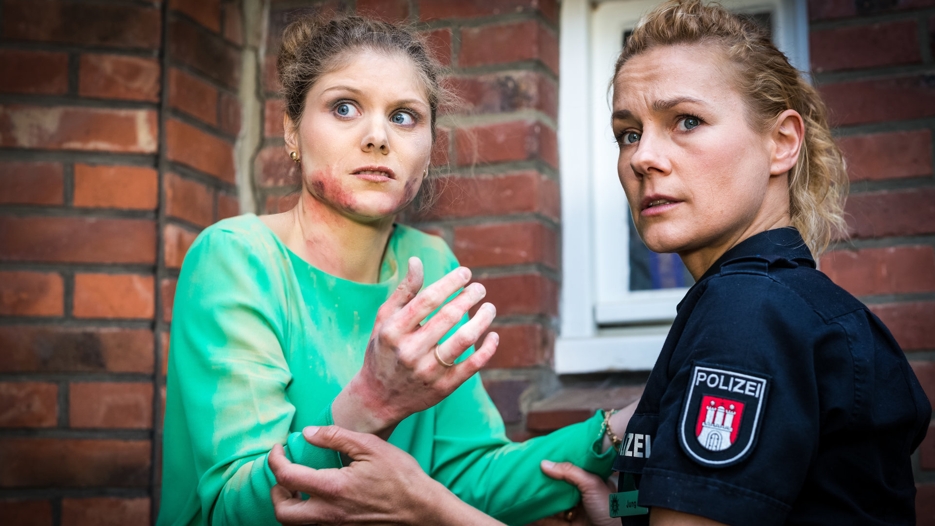 "Notruf Hafenkante - Notwehr": Valerie Politz (Stephanie Lexer) steht vor einem Backsteinhaus und sieht erschrocken zur Seite. Sie hat Verletzungen im Gesicht und an den Händen, die sie vor sich hält. Die Polizistin Franzi Jung (Rhea Harder-Vennewald) steht vor ihr und hält beruhigend ihre Unterarme. Franzi schaut in die gleiche Richtung wie Valerie.