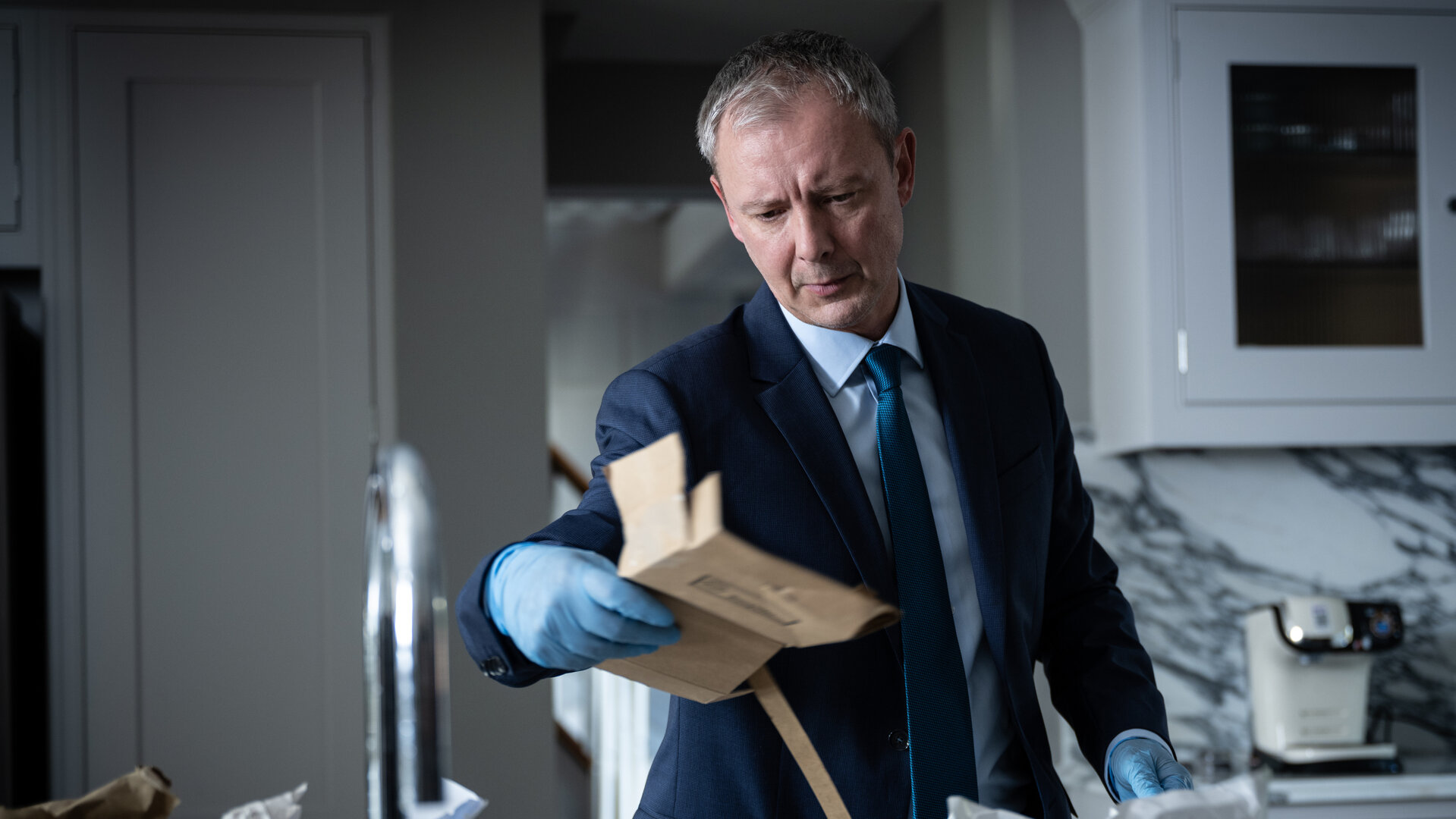 "Detective Grace - Nicht tot genug": DS Roy Grace (John Simm) steht in einer Küche und untersucht ein Stück Pappverpackung, das er in der linken Hand hält. Er trägt blaue Einweg-Handschuhe.