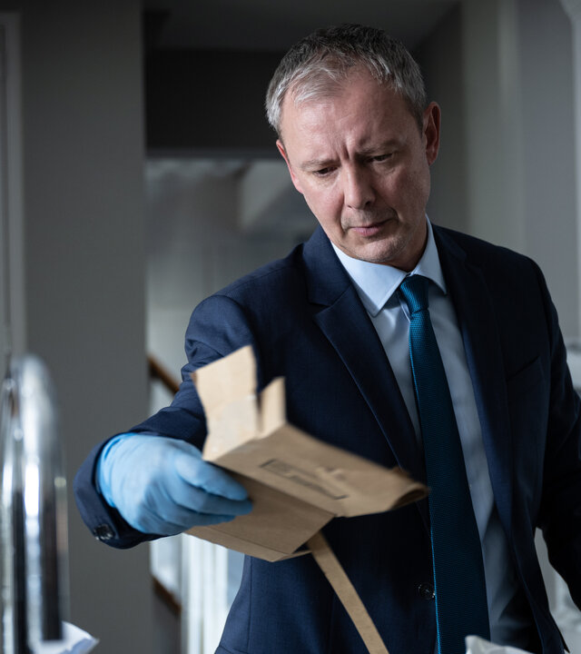 "Detective Grace - Nicht tot genug": DS Roy Grace (John Simm) steht in einer Küche und untersucht ein Stück Pappverpackung, das er in der linken Hand hält. Er trägt blaue Einweg-Handschuhe.