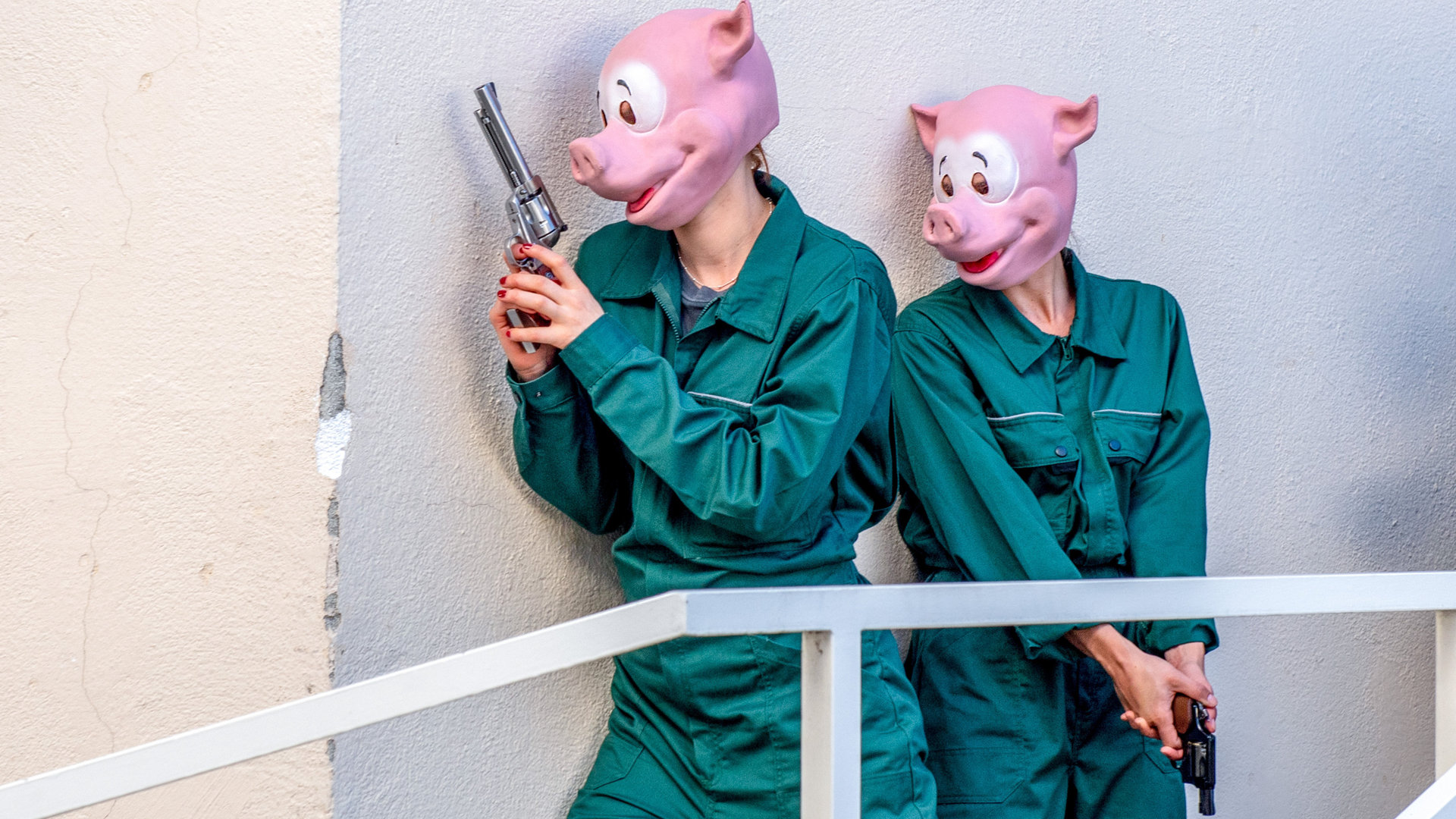 "SOKO Kitzbühel - Auf der Flucht": Zwei Frauen in grünen Overalls und Schweinemasken lehnen sich bewaffnet an eine Wand und blicken in dieselbe Richtung.