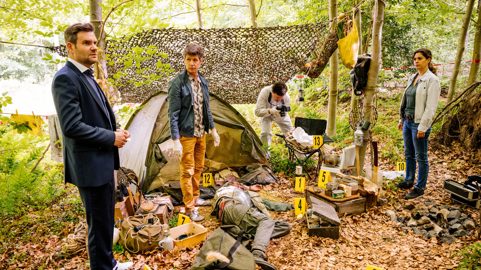 "SOKO Köln - Fuchsmann": Pathologe Dr. Kraft (Thomas Clemens) steht mit Jonas Fischer (Lukas Piloty) und Lilly Funke (Tatjana Kästel) an einem Tatort in einem Wald. Zwischen ihnen liegt eine Leiche vor einem Zelt. Auf dem mit Laub bedeckten Boden liegen diverse Taschen, Koffer und Klamotten verstreut, außerdem stehen viele gelbe Plastikkarten mit Zahlen dort. Ein Mitarbeiter der Spurensicherung (Komparse) fotografiert einen Gegenstand auf einem Stuhl. Im Hintergrund sieht man eine braune Hängematte zwischen Bäumen befestigt schweben.