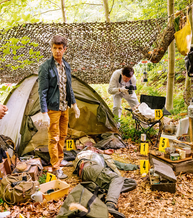 "SOKO Köln - Fuchsmann": Pathologe Dr. Kraft (Thomas Clemens) steht mit Jonas Fischer (Lukas Piloty) und Lilly Funke (Tatjana Kästel) an einem Tatort in einem Wald. Zwischen ihnen liegt eine Leiche vor einem Zelt. Auf dem mit Laub bedeckten Boden liegen diverse Taschen, Koffer und Klamotten verstreut, außerdem stehen viele gelbe Plastikkarten mit Zahlen dort. Ein Mitarbeiter der Spurensicherung (Komparse) fotografiert einen Gegenstand auf einem Stuhl. Im Hintergrund sieht man eine braune Hängematte zwischen Bäumen befestigt schweben.