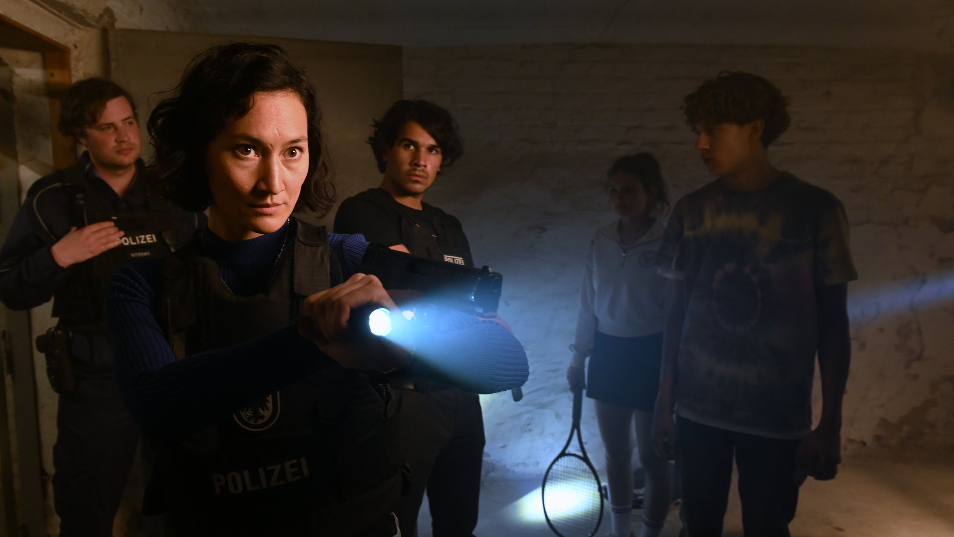 "SOKO Potsdam - Romy und Julian": In einem Kellerraum steht Pauline Hobrecht (Agnes Decker) mit kugelsicherer Weste. Sie hat eine Taschenlampe und und ihre Dienstwaffe erhoben und auf etwas außerhalb des Bildes gerichtet. Samir Amari (Skandar Amini) steht ein paar Schritte hinter ihr und sieht sie aufmerksam an. Rechts neben ihm stehen Romy Lubitz (Greta Bohacek), die einen Tennisschläger in der Hand hat, und Julian Schmitt (Marven Gabriel Suarez-Brinkert). Links im Bild steht ein weiterer Polizist (Komparse), der den Blick auf Julian gerichtet hat.