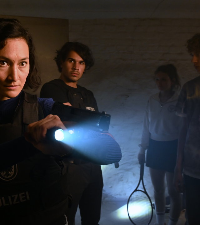 "SOKO Potsdam - Romy und Julian": In einem Kellerraum steht Pauline Hobrecht (Agnes Decker) mit kugelsicherer Weste. Sie hat eine Taschenlampe und und ihre Dienstwaffe erhoben und auf etwas außerhalb des Bildes gerichtet. Samir Amari (Skandar Amini) steht ein paar Schritte hinter ihr und sieht sie aufmerksam an. Rechts neben ihm stehen Romy Lubitz (Greta Bohacek), die einen Tennisschläger in der Hand hat, und Julian Schmitt (Marven Gabriel Suarez-Brinkert). Links im Bild steht ein weiterer Polizist (Komparse), der den Blick auf Julian gerichtet hat.