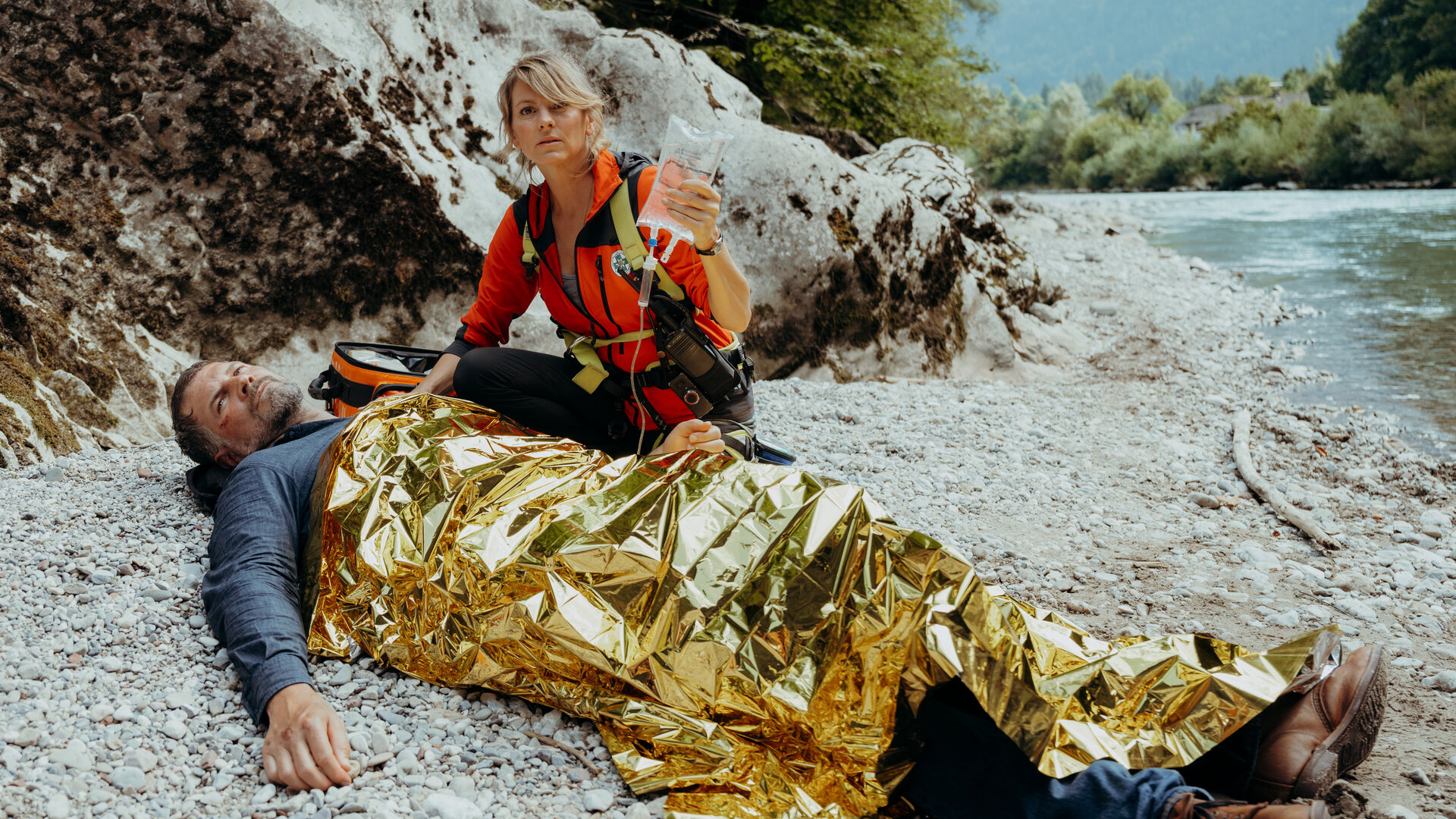 "Die Bergretter - Aus Angst (2)": Am steinigen Ufer eines Bachlaufs: In eine Rettungsdecke eingewickelt liegt Robert Eichner (Simon Böer) am Boden. Neben ihm kniet Bergretterin Katharina (Luise Bähr) und hält eine Infusion in der Hand.