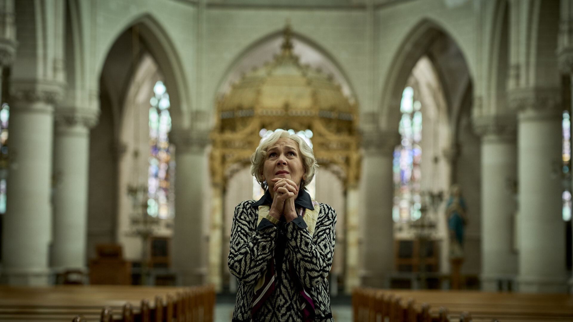 "Elixir – The Pain Killers - Die Familie Rombauts": Riekje Rombauts (Katelijne Damen) steht im Mittelgang einer Kirche. Sie faltet ihre Hände und schaut nach oben. Ihr Gesichtsausdruck ist besorgt.