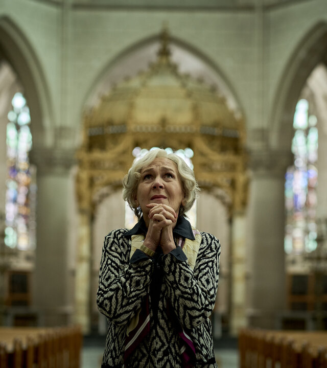 "Elixir – The Pain Killers - Die Familie Rombauts": Riekje Rombauts (Katelijne Damen) steht im Mittelgang einer Kirche. Sie faltet ihre Hände und schaut nach oben. Ihr Gesichtsausdruck ist besorgt.