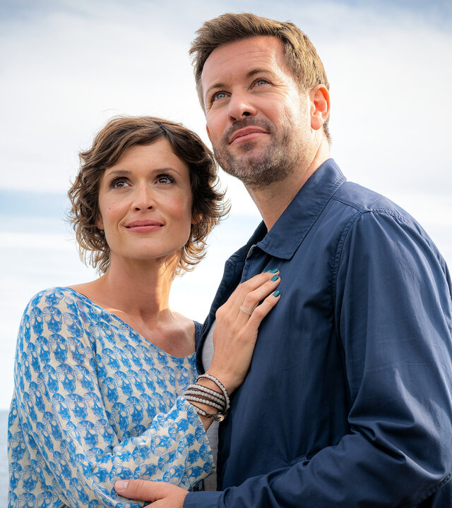 "Inga Lindström: Sag' einfach ja": Am Ufer: Linda (Sophie Melbinger) und Tomas (Jan Hartmann) halten sich in den Armen und schauen glücklich in die Ferne. Linda hat ihre Hand auf Tomas' Brust gelegt, während er ihren Arm zart berührt.