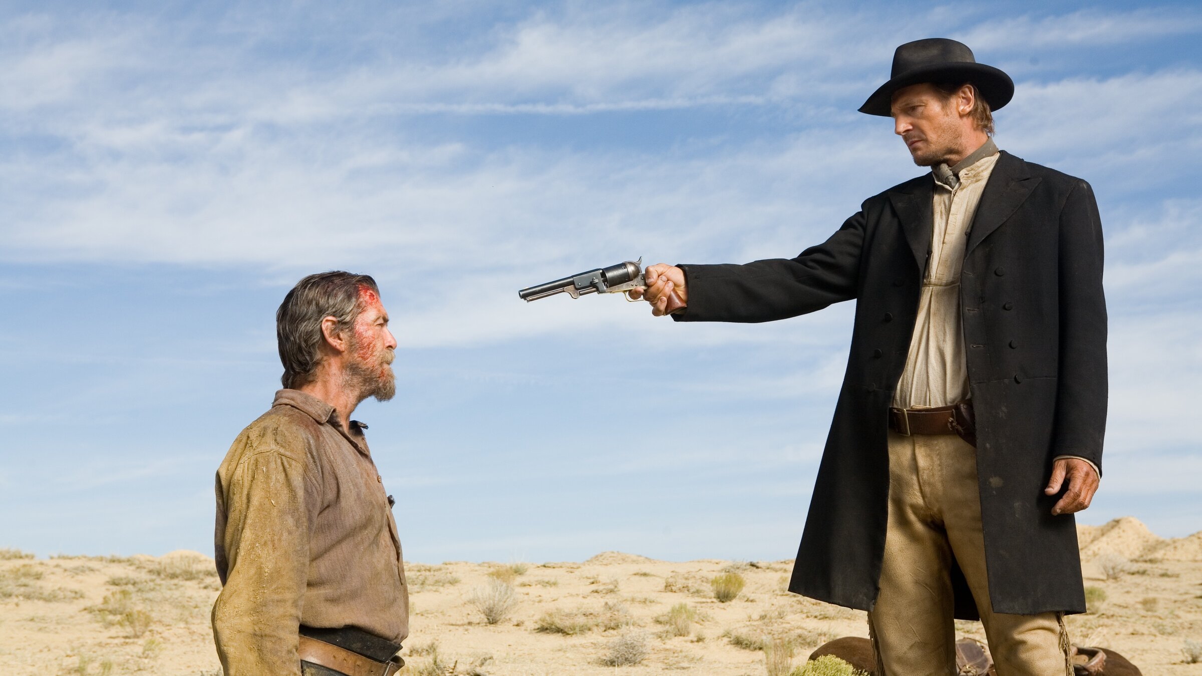 „Seraphim Falls – Gnadenlose Jagd“: Carver (Liam Neeson, rechts), in langem schwarzen Mantel und Hut, zielt mit einem Revolver aus nächster Nähe auf den Kopf von Gideon (Pierce Brosnan), der vor ihm kniet. Im Hintergrund ist eine wüstenähnliche Landschaft zu sehen.