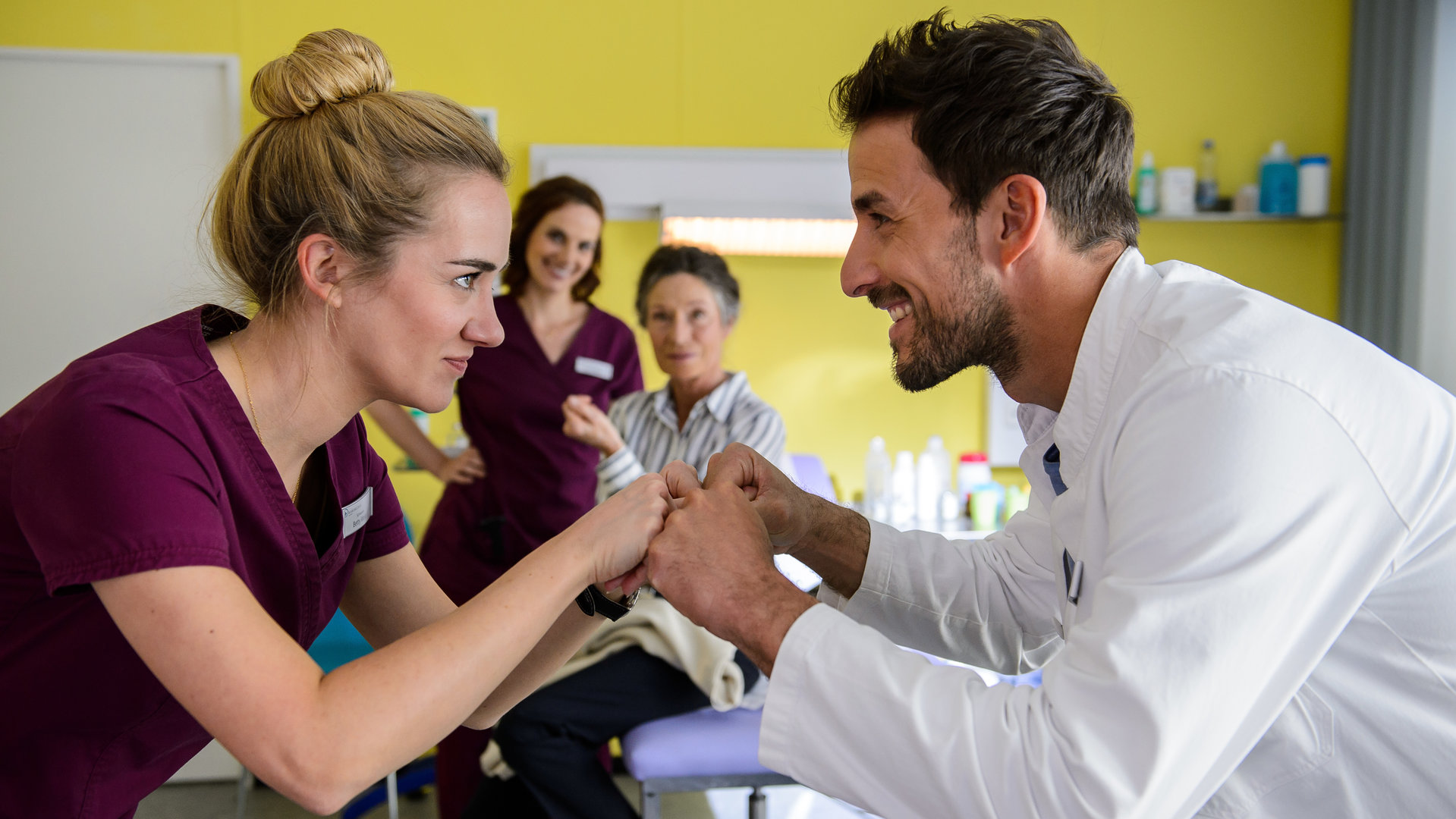 "Bettys Diagnose - Eins mit Sternchen": Betty Weiss (Annina Hellenthal) und Dr. Frank Stern (Max Alberti) halten ihre Fäuste aneinander, Hanna Winter (Marie Zielcke) und Dagmar Schönemann (Renate Serwotke) befinden sich im Hintergrund.