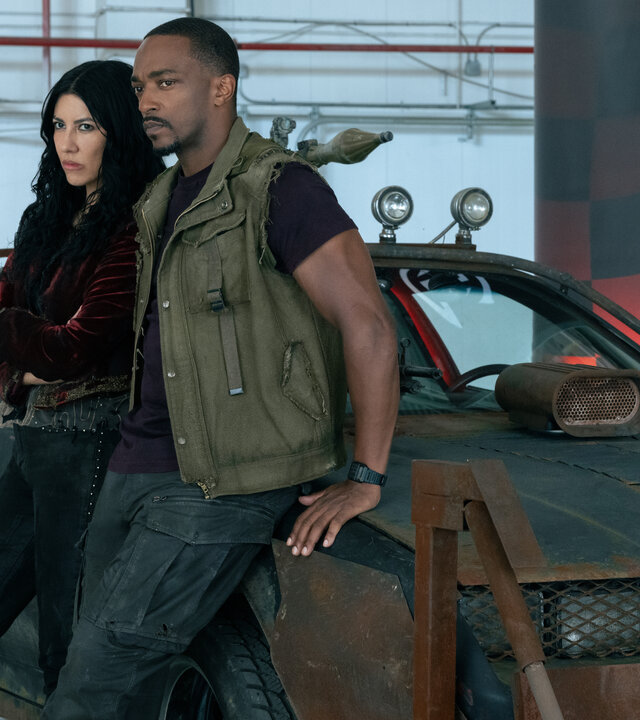 "Twisted Metal - ONURMRK": In einer Halle stehen Quiet (Stephanie Beatriz) und John (Anthony Mackie) vor einem sportlichen, kampfbereit wirkenden Fahrzeug. Beide lehnen an der Motorhaube und blicken mit ernster Miene zur Seite. John hat seine Hände hinter hinter sich auf die Motorhaube gelegt. Das Fahrzeug weist mehrere braune Rostflecken auf und ist mit auffälligen Modifikationen wie zusätzlichen Scheinwerfern, Waffen und dekorativen Elementen am Kühlergrill ausgestattet.