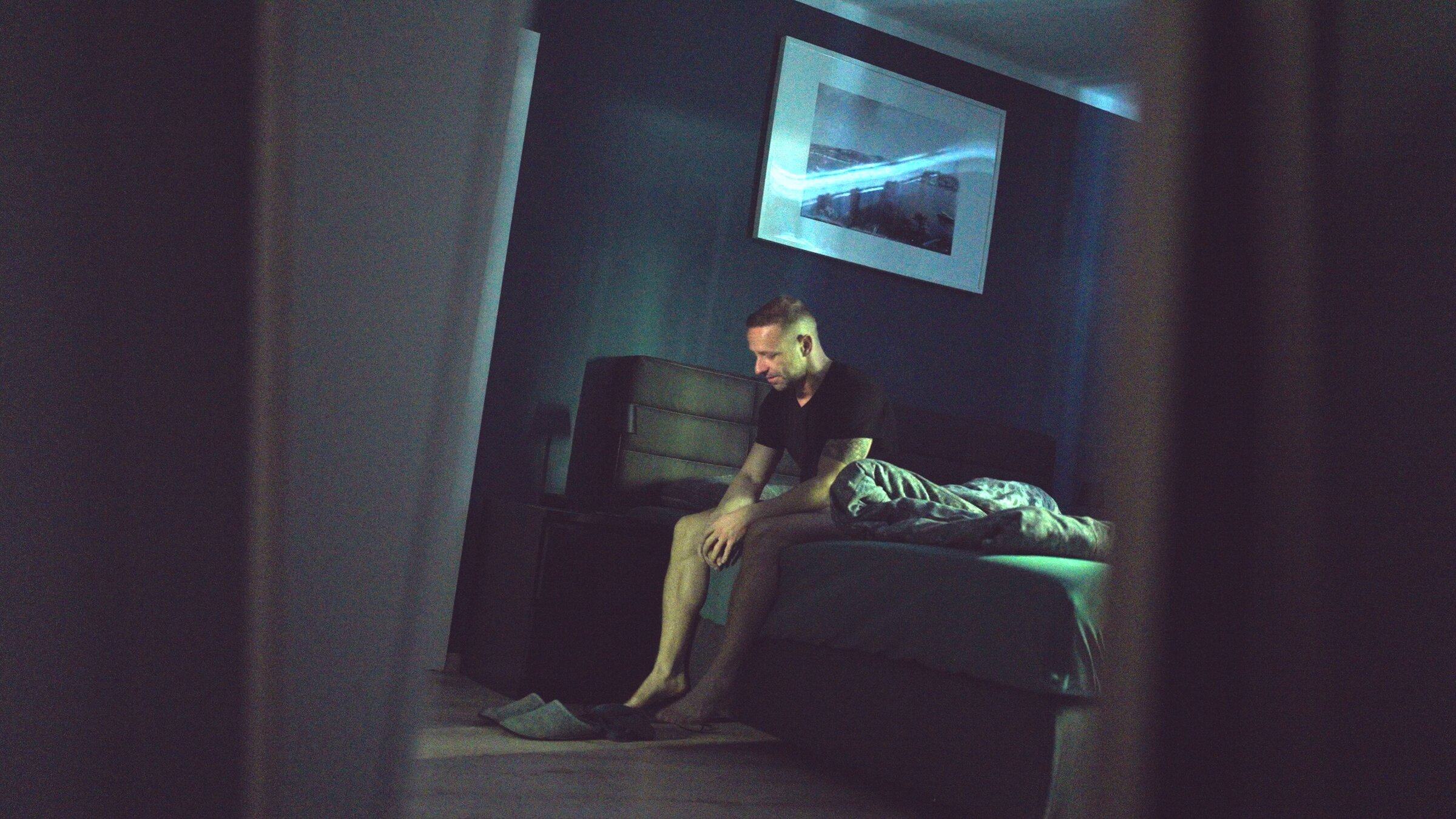 "37°: Schlaflos: Wenn die Nacht zum Feind wird": Der 36-Jährige Waldemar sitzt auf der Bettkante im dunklen Schlafzimmer und schaut nach unten. Über ihm hängt ein Bild an der Wand.