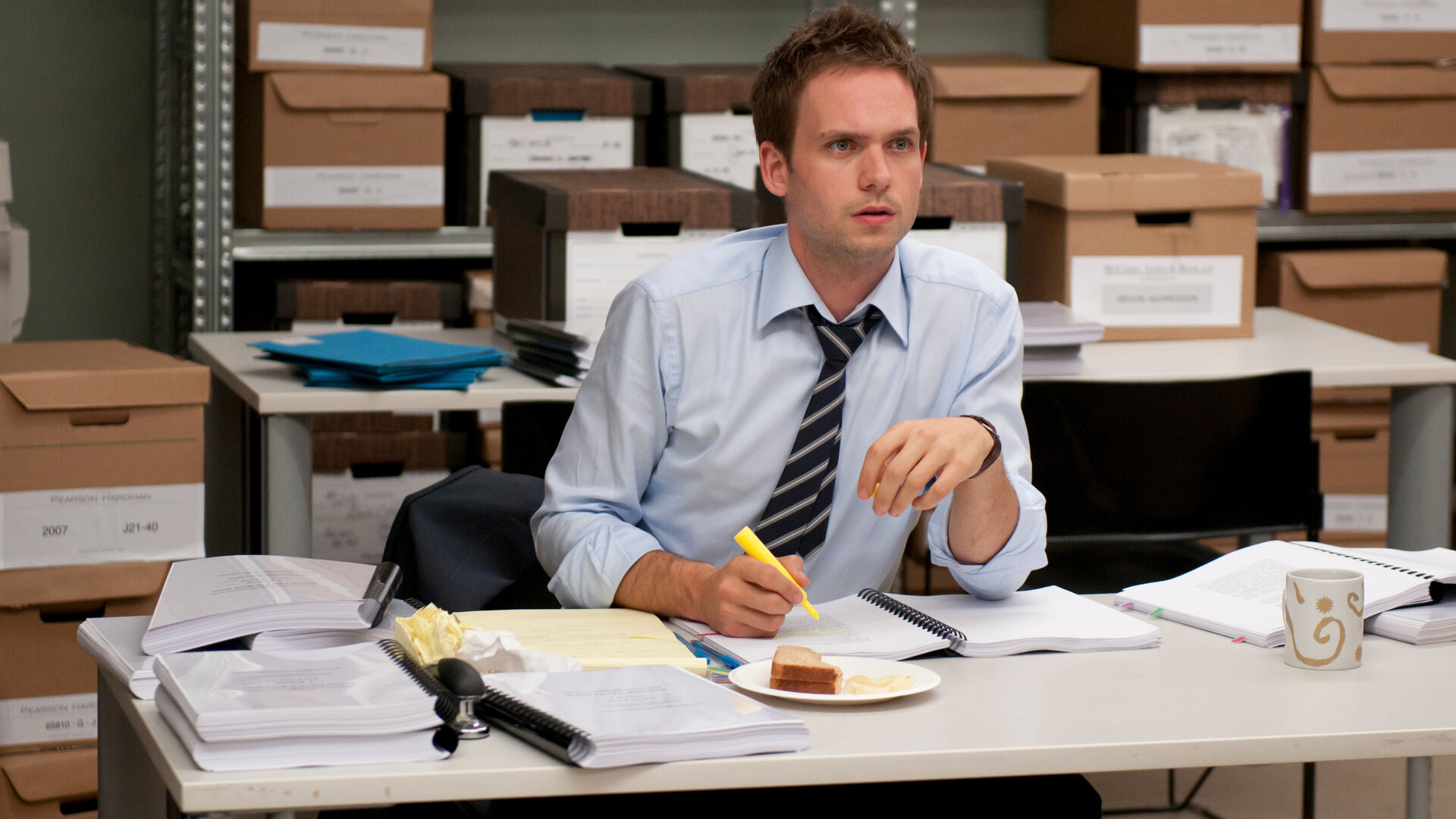 "Suits - Patente und andere Unwägbarkeiten": Mike Ross (Patrick J. Adams) sitzt mit hochgekrempelten Hemdsärmeln an einem überfüllten Tisch im Archiv, in der Hand hält er einen Textmarker. Vor ihm liegen zahlreiche Akten, daneben steht eine Tasse und ein Teller mit einem halb gegessenen Sandwich; im Hintergrund sind Kartons auf Metallregalen gestapelt.