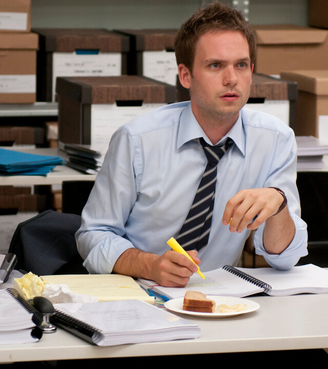 "Suits - Patente und andere Unwägbarkeiten": Mike Ross (Patrick J. Adams) sitzt mit hochgekrempelten Hemdsärmeln an einem überfüllten Tisch im Archiv, in der Hand hält er einen Textmarker. Vor ihm liegen zahlreiche Akten, daneben steht eine Tasse und ein Teller mit einem halb gegessenen Sandwich; im Hintergrund sind Kartons auf Metallregalen gestapelt.