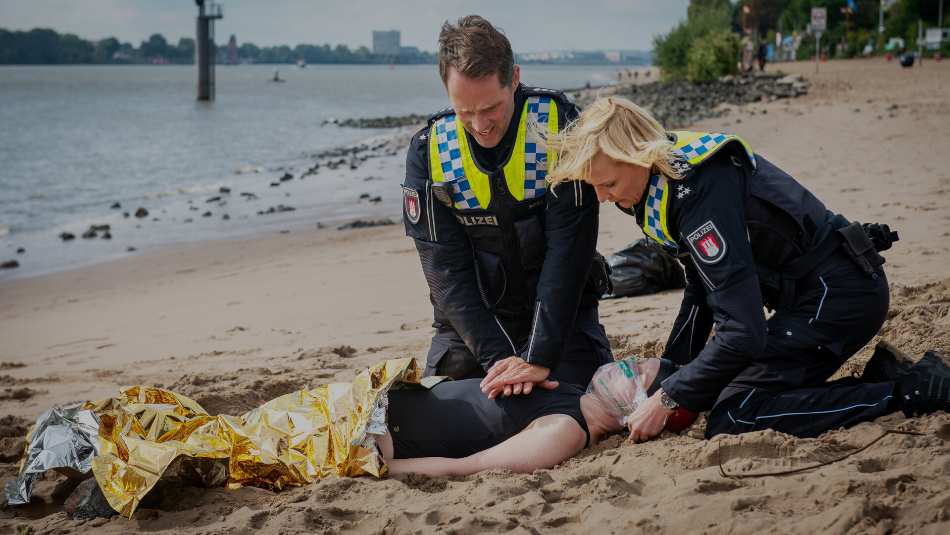 "Notruf Hafenkante - Eiskalt": Am Elbstrand. Der Polizist Mattes Seeler (Matthias Schloo) und seine Kollegin Melanie Hansen (Sanna Englund) knien auf dem Sand und sind dabei Erste-Hilfe-Maßnahmen an einer Frau auszuführen, die rücklings auf dem Boden liegt. Eine Wärmedecke liegt über ihrem Körper.