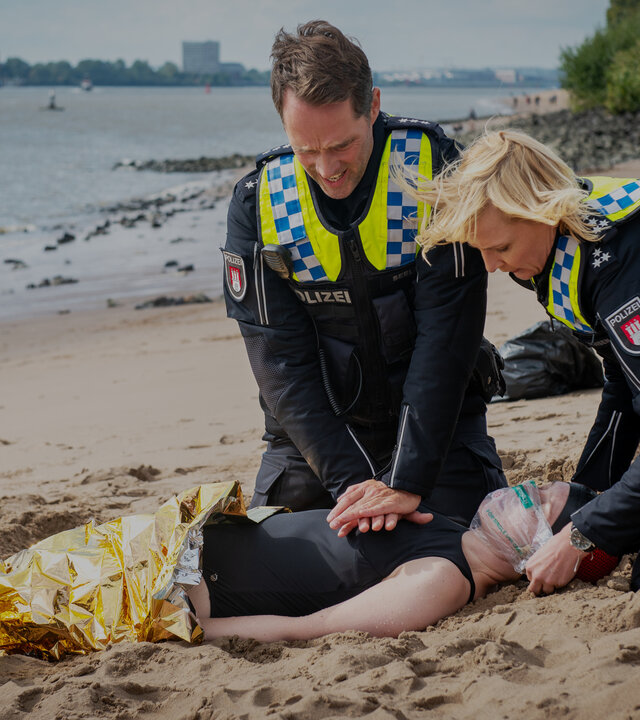 "Notruf Hafenkante - Eiskalt": Am Elbstrand. Der Polizist Mattes Seeler (Matthias Schloo) und seine Kollegin Melanie Hansen (Sanna Englund) knien auf dem Sand und sind dabei Erste-Hilfe-Maßnahmen an einer Frau auszuführen, die rücklings auf dem Boden liegt. Eine Wärmedecke liegt über ihrem Körper.