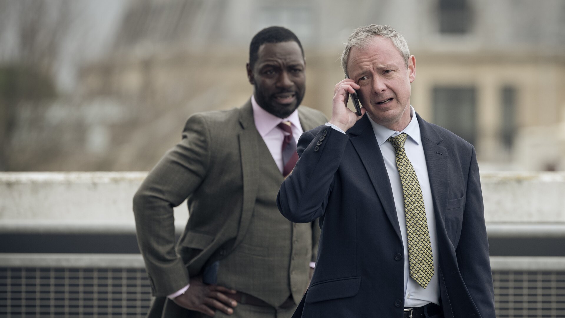 "Detective Grace - Die Zeit läuft": DS Roy Grace (John Simm) und DS Glenn Branson (Richie Campbell) stehen nebeneinander vor einem Gebäude, das nur unscharf im Hintergrund zu erkennen ist. Grace hält sein Handy ans rechte Ohr und telefoniert. Branson hat seine Arme in die Hüften gestemmt und schaut Grace an.