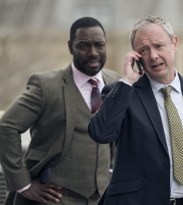 "Detective Grace - Die Zeit läuft": DS Roy Grace (John Simm) und DS Glenn Branson (Richie Campbell) stehen nebeneinander vor einem Gebäude, das nur unscharf im Hintergrund zu erkennen ist. Grace hält sein Handy ans rechte Ohr und telefoniert. Branson hat seine Arme in die Hüften gestemmt und schaut Grace an.