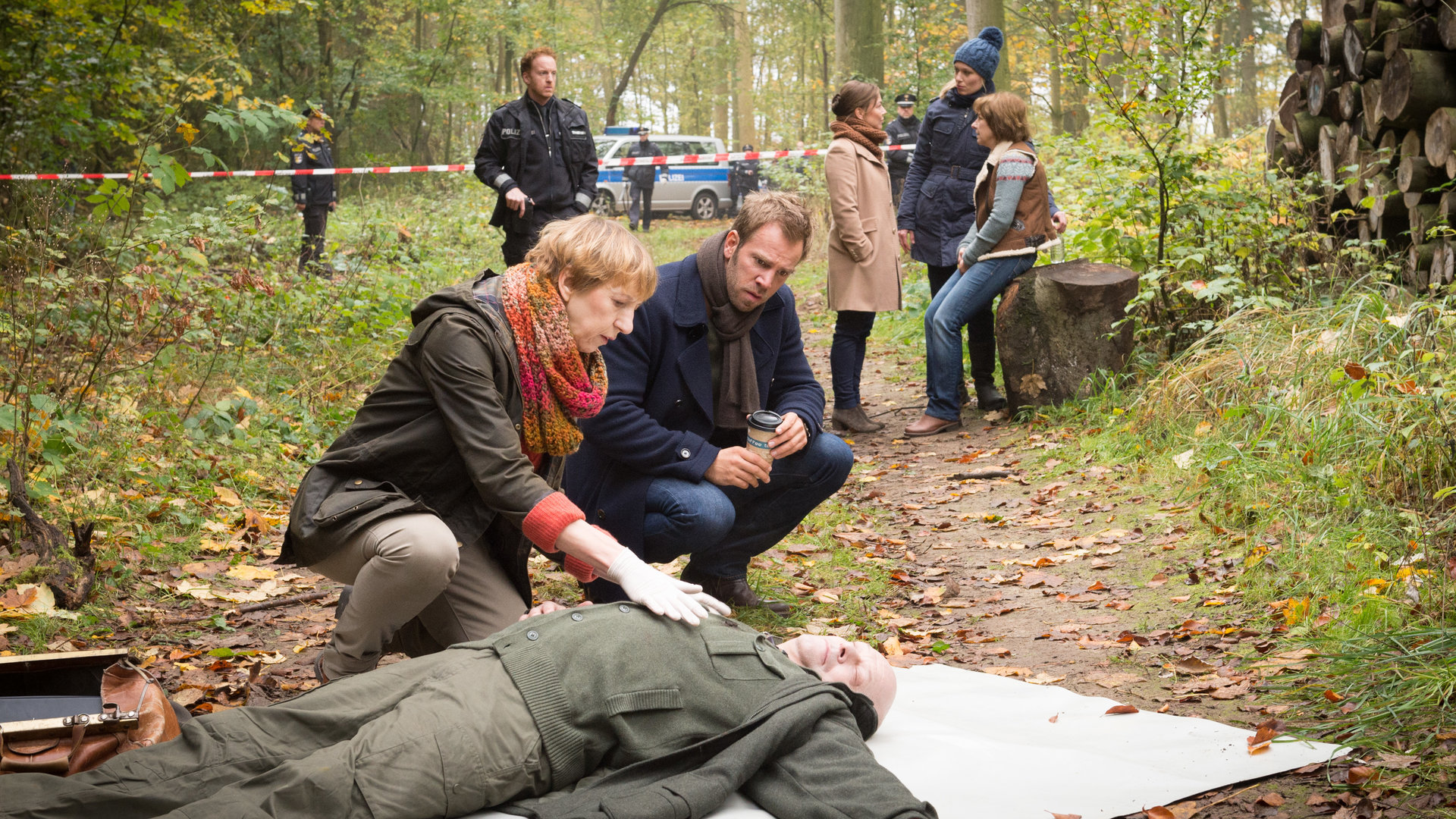 „SOKO Wismar – Der letzte Zeuge“: Der tote Hans Berger (Harald Weiler) liegt auf dem Waldboden. Pathologin Helene Sturbeck (Katharina Blaschke) tastet den Toten ab. Kommissar Lars Pöhlmann (Dominic Boeer) kniet neben ihr und schaut aufmerksam dabei zu. Im Hintergrund stehen Timmermann (Mathias Junge) sowie Börensen (Claudia Schmutzler) und van der Meer (Isabel Berghout), die mit der Schwester des Toten, Ute Berger (Irene Rindje), reden.