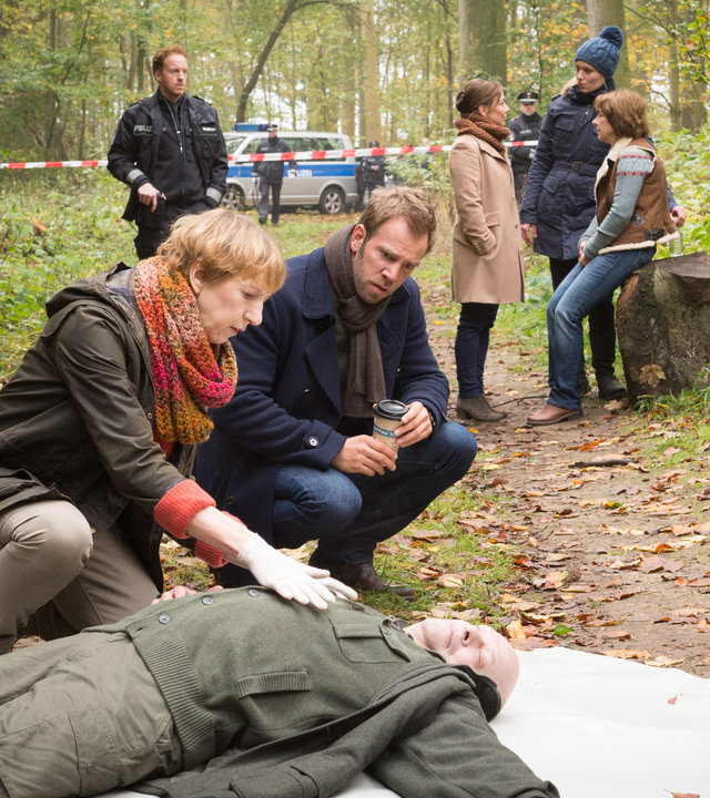„SOKO Wismar – Der letzte Zeuge“: Der tote Hans Berger (Harald Weiler) liegt auf dem Waldboden. Pathologin Helene Sturbeck (Katharina Blaschke) tastet den Toten ab. Kommissar Lars Pöhlmann (Dominic Boeer) kniet neben ihr und schaut aufmerksam dabei zu. Im Hintergrund stehen Timmermann (Mathias Junge) sowie Börensen (Claudia Schmutzler) und van der Meer (Isabel Berghout), die mit der Schwester des Toten, Ute Berger (Irene Rindje), reden.
