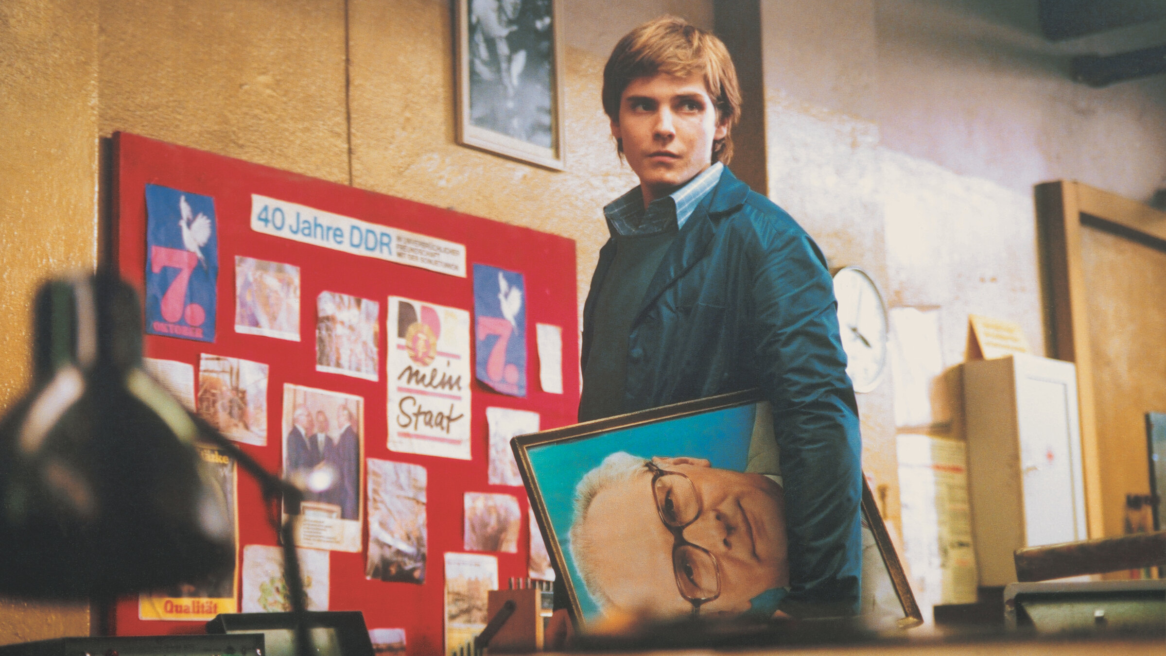 „Good bye, Lenin!“: Alex (Daniel Brühl) blickt in einer Art Lagerhalle suchend um sich. Unter dem linken Arm trägt er ein Porträt Erich Honeckers. Im Hintergrund sieht man eine Fototafel mit der Überschrift „40 Jahre DDR“.
