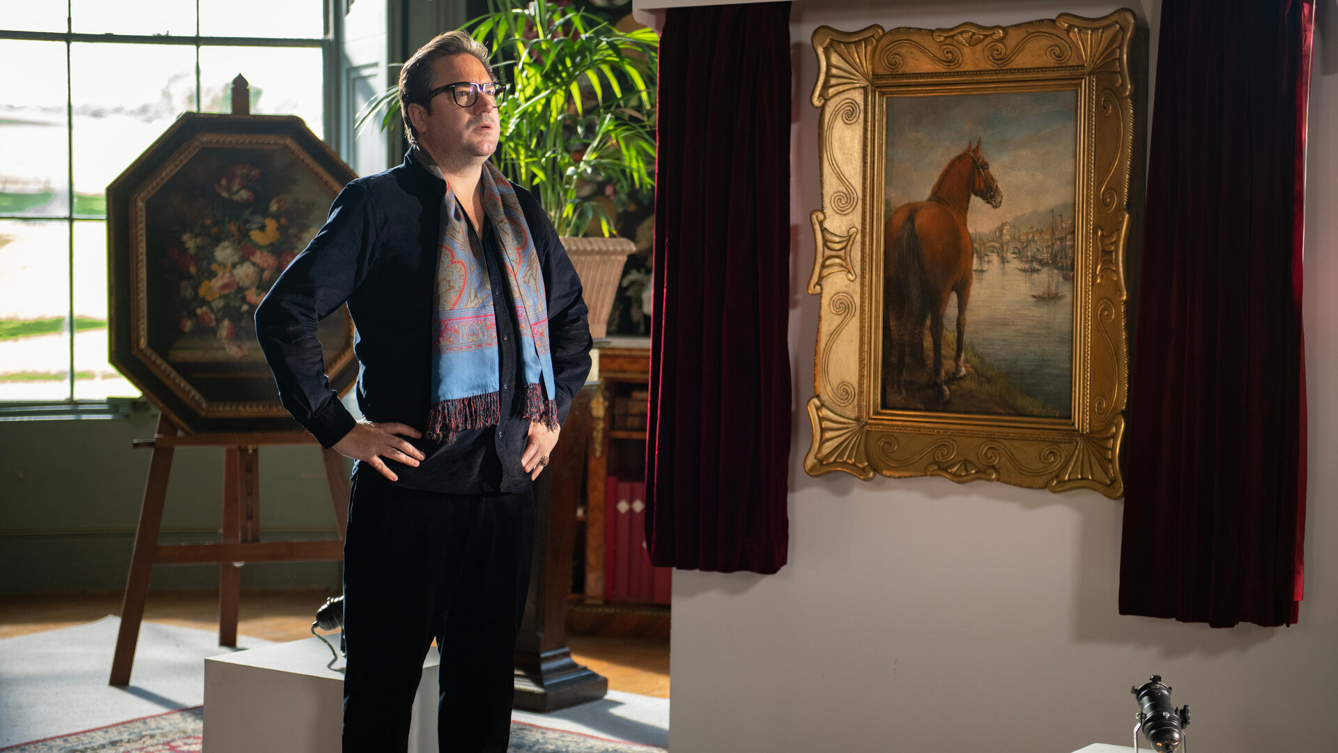 "Beyond Paradise - Der Kunstraub": Kunsthändler Terrence Witham (Rufus Jones) steht neben dem Gemälde "Solo Mare", auf dem ein Pferd zu sehen ist. Er ist schwarz gekleidet und trägt einen auffällig bunten Schal.