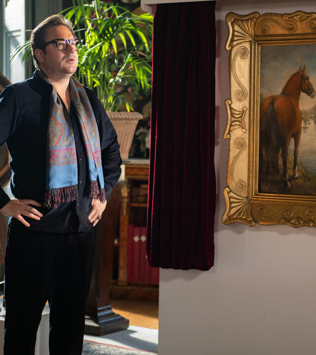 "Beyond Paradise - Der Kunstraub": Kunsthändler Terrence Witham (Rufus Jones) steht neben dem Gemälde "Solo Mare", auf dem ein Pferd zu sehen ist. Er ist schwarz gekleidet und trägt einen auffällig bunten Schal.