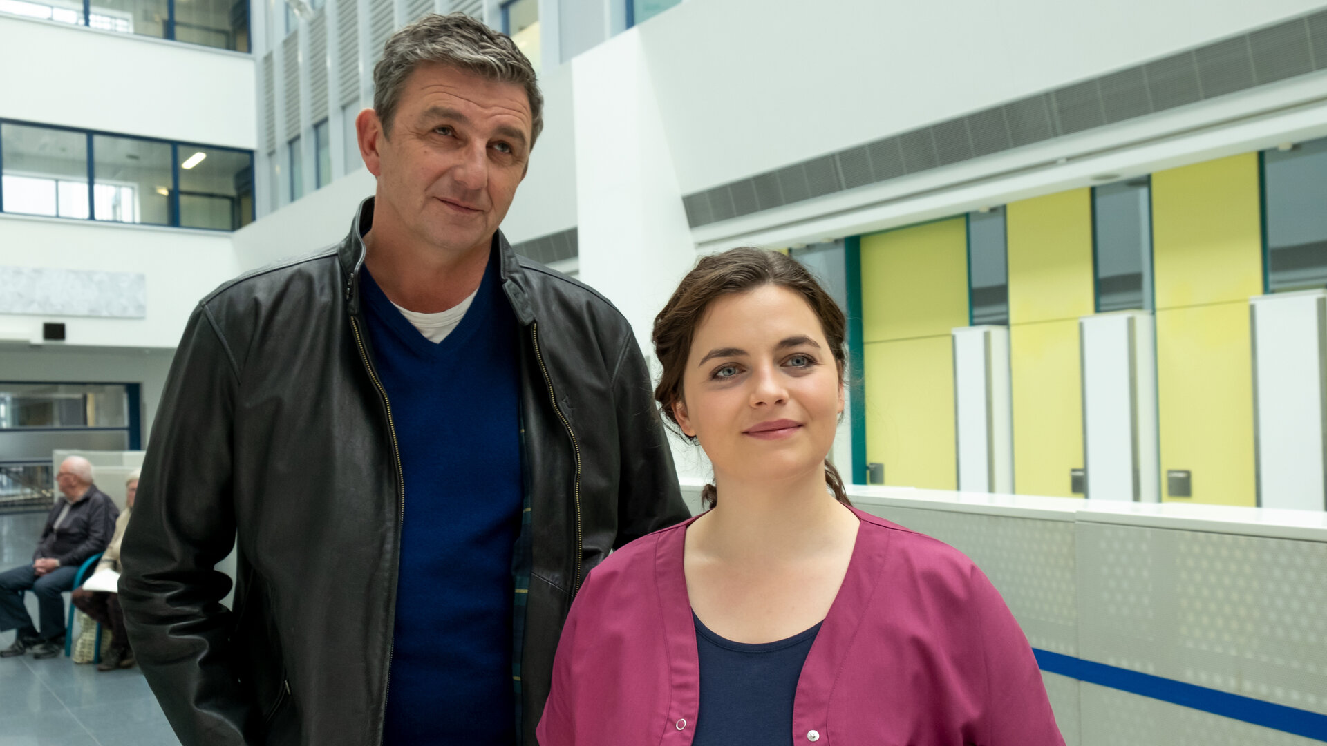 "Der Bergdoktor: Neuland (2)": Im Krankenhaus: Martin (Hans Sigl) steht, die Hände in den Hosentaschen, hinter Lilli (Ronja Forcher), die ihre Arbeitskleidung trägt. Beide blicken in die Ferne und lächeln.