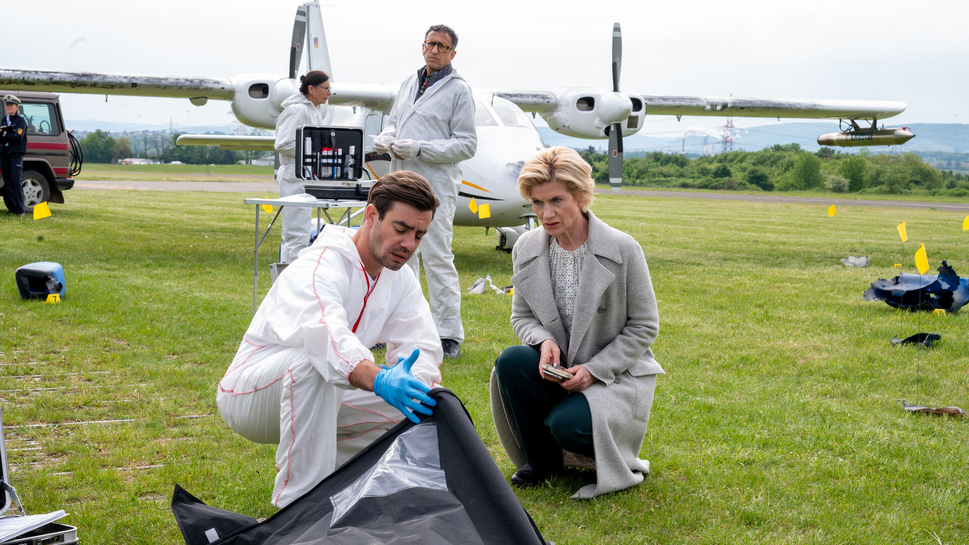 "SOKO Stuttgart - Hagelflieger": Die Erste Kriminalhauptkommissarin Martina Seiffert (Astrid M. Fünderich) und der Leiter der Rechtsmedizin Dr. Benedikt Förster (Florian Wünsche) knien auf einem Flughafengelände am Boden. Ihr Blick fällt in einen schwarzen Leichensack, den Dr. Förster aufhält: Im Hintergrund steht der Leiter der KTU, Jan Arnaud (Mike Zaka Sommerfeldt), vor einer Propellermaschine und schaut zu den beiden. Daneben arbeitet eine Kollegin (Komparsin) der Spurensicherung.