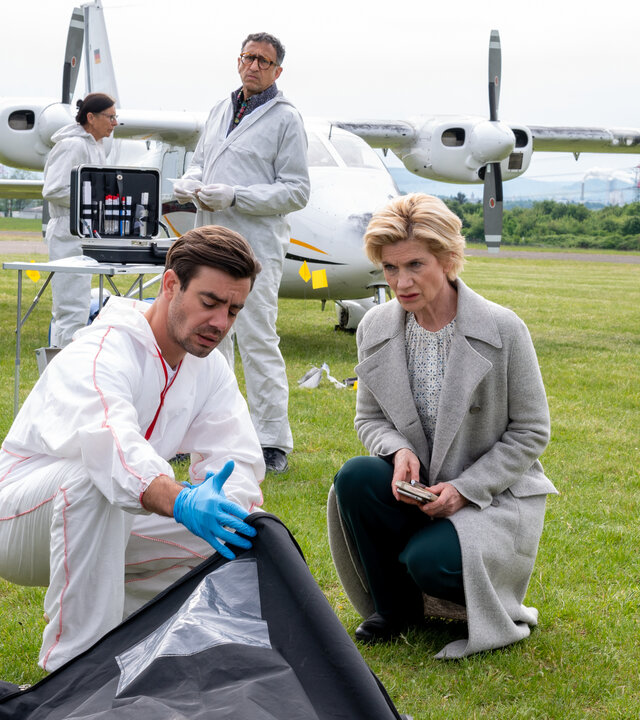 "SOKO Stuttgart - Hagelflieger": Die Erste Kriminalhauptkommissarin Martina Seiffert (Astrid M. Fünderich) und der Leiter der Rechtsmedizin Dr. Benedikt Förster (Florian Wünsche) knien auf einem Flughafengelände am Boden. Ihr Blick fällt in einen schwarzen Leichensack, den Dr. Förster aufhält: Im Hintergrund steht der Leiter der KTU, Jan Arnaud (Mike Zaka Sommerfeldt), vor einer Propellermaschine und schaut zu den beiden. Daneben arbeitet eine Kollegin (Komparsin) der Spurensicherung.