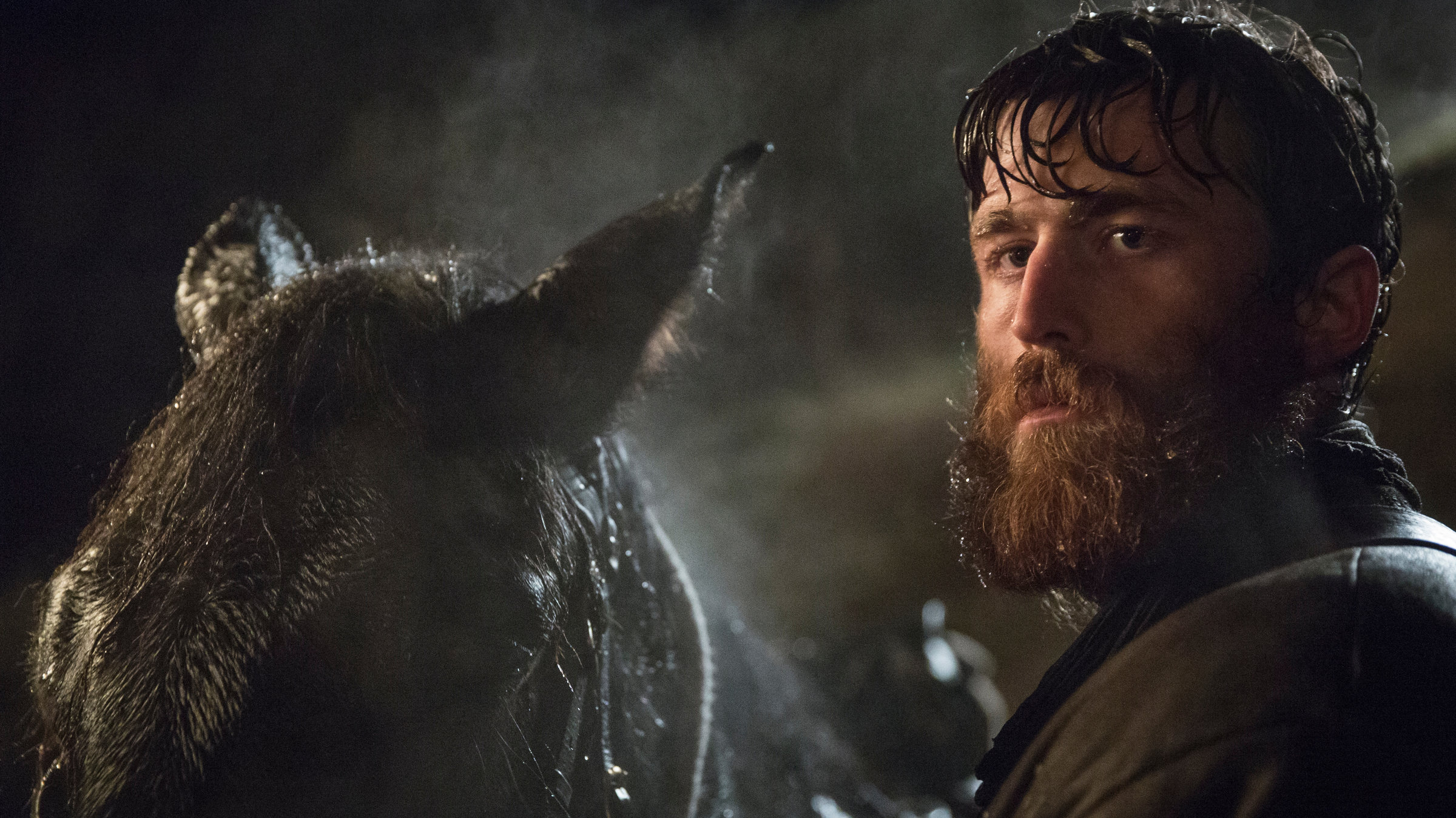 "Black47": Ein wild aussehender Mann mit rötlichem Vollbart steht in der Nacht in feuchtem Wetter neben einem Pferd und blickt aufmerksam um sich.