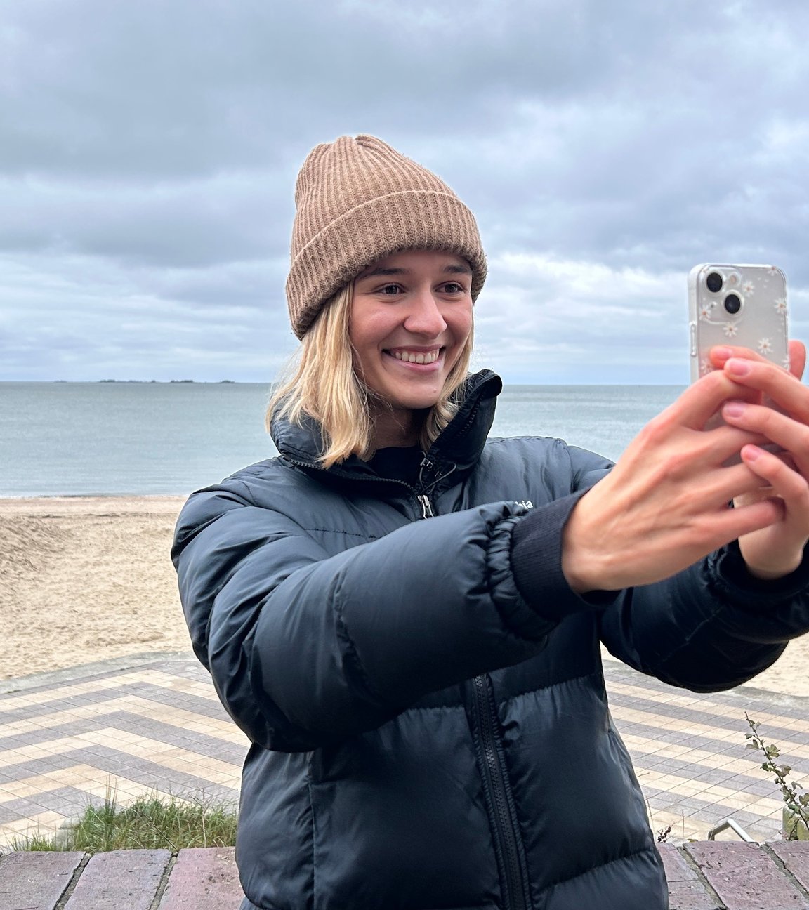 "ZDF.reportage: Winter am Wattenmeer - Einsame Inseln": Ariana Müller steht auf der Strandpromenade, hinter ihr liegt der leere Strand und das Meer. Sie nimmt gerade ein Video mit dem Smartphone auf.