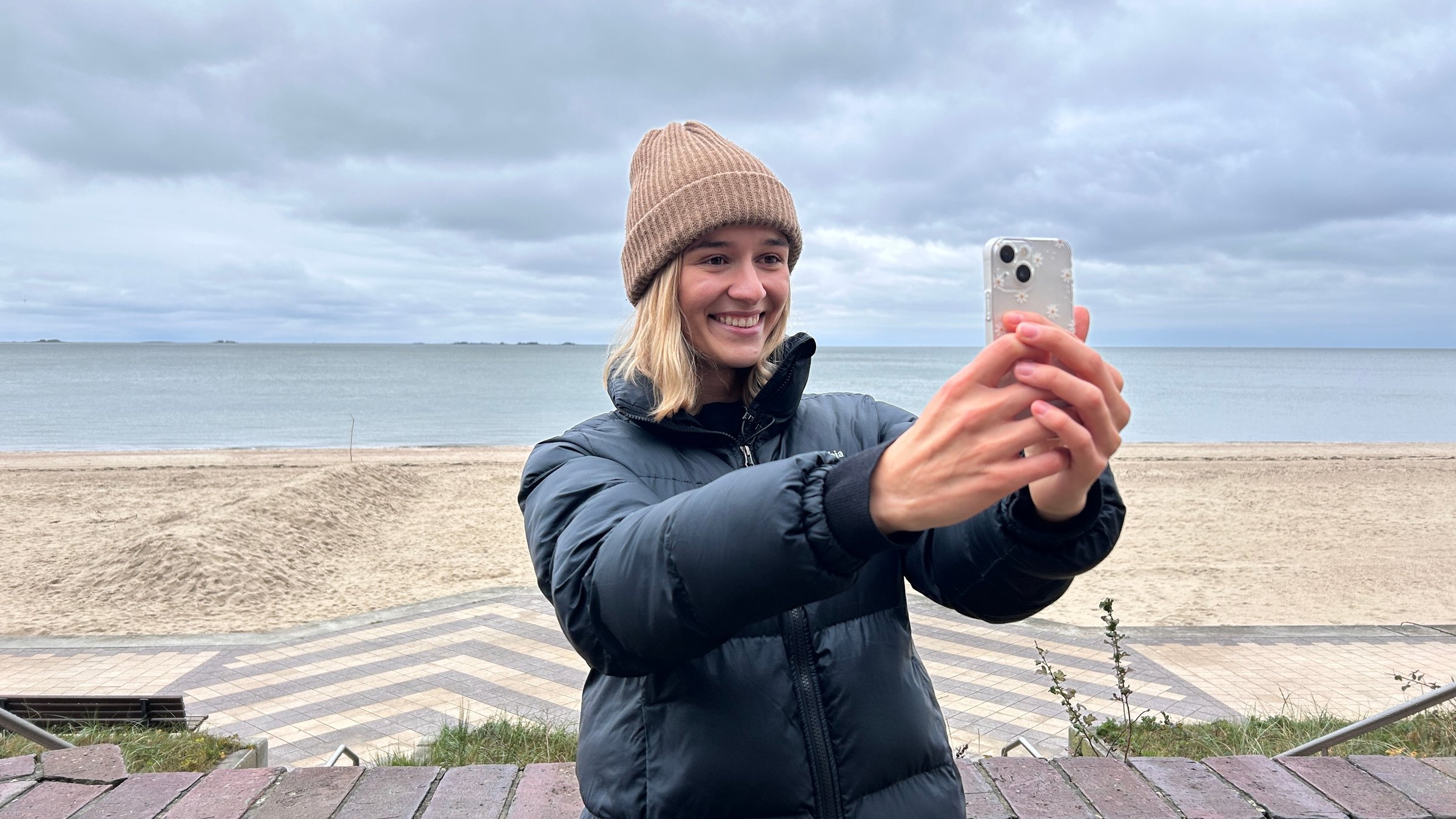 "ZDF.reportage: Winter am Wattenmeer - Einsame Inseln": Ariana Müller steht auf der Strandpromenade, hinter ihr liegt der leere Strand und das Meer. Sie nimmt gerade ein Video mit dem Smartphone auf.