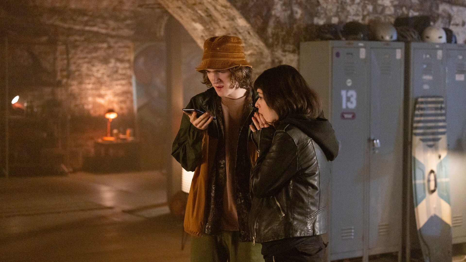 "Alex Rider - Feind": Tom (Brenock O'Connor) steht in einem düsteren Raum mit Steinwänden und hält ein Smartphone in der Hand. Er scheint mit jemandem zu sprechen, während Kyra (Marli Siu) neben ihm steht und sich schockiert die Hände vor den Mund hält. Hinter ihnen steht ein Spind, an dem ein Wakeboard lehnt.