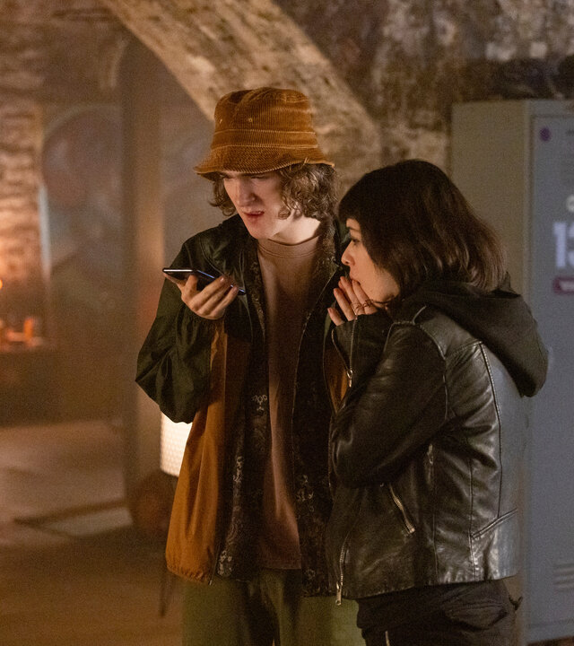 "Alex Rider - Feind": Tom (Brenock O'Connor) steht in einem düsteren Raum mit Steinwänden und hält ein Smartphone in der Hand. Er scheint mit jemandem zu sprechen, während Kyra (Marli Siu) neben ihm steht und sich schockiert die Hände vor den Mund hält. Hinter ihnen steht ein Spind, an dem ein Wakeboard lehnt.