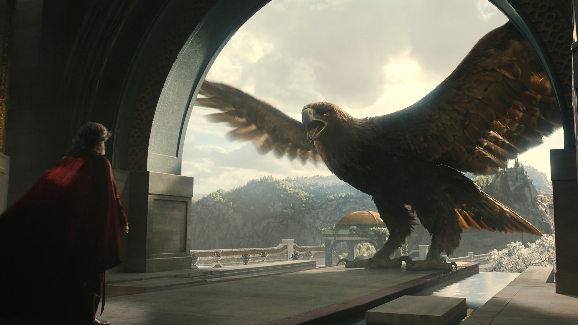 "Der Herr der Ringe: Die Ringe der Macht - Der Adler und das Zepter": Ein riesiger Adler steht mit ausgebreiteten Schwingen in einem Torbogen. Hinter ihm sind draußen die Hügel von Númenor zu erkennen. Dem Adler gegenüber steht Ar-Pharazôn (Trystan Gravelle) in einem Saal. Er ist nur von hinten zu sehen.